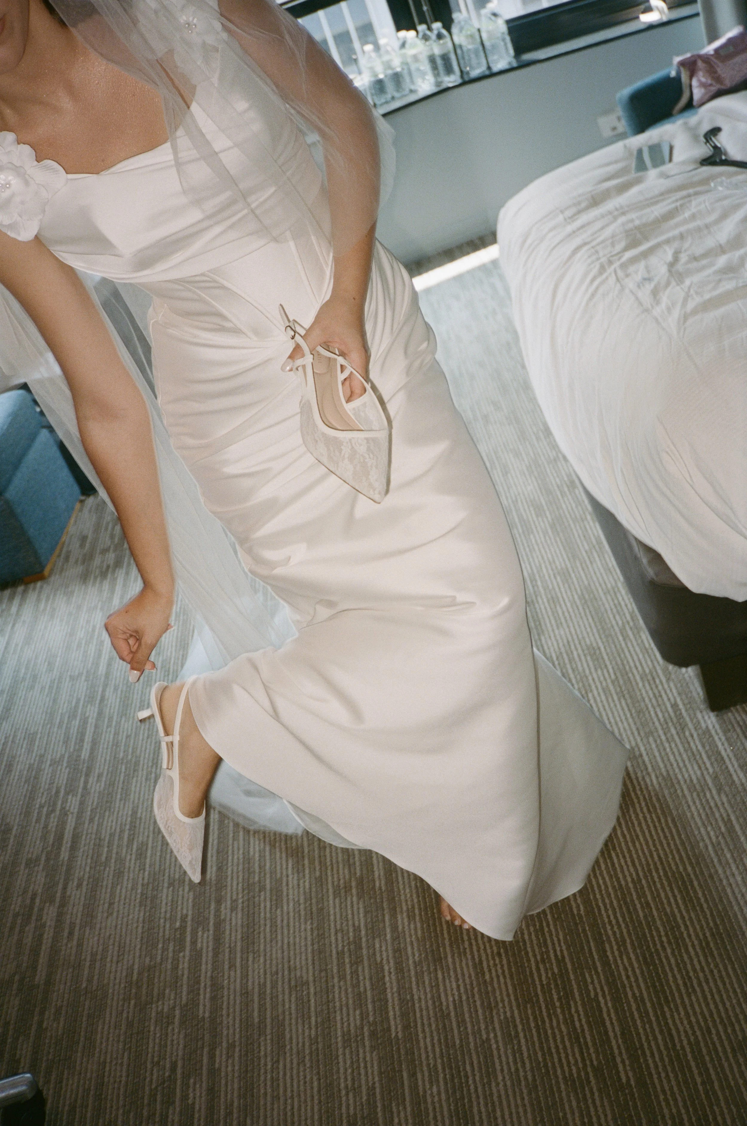 Person in a white wedding dress holding a lace high heel shoe and lifting one foot, possibly preparing to put on the shoe.