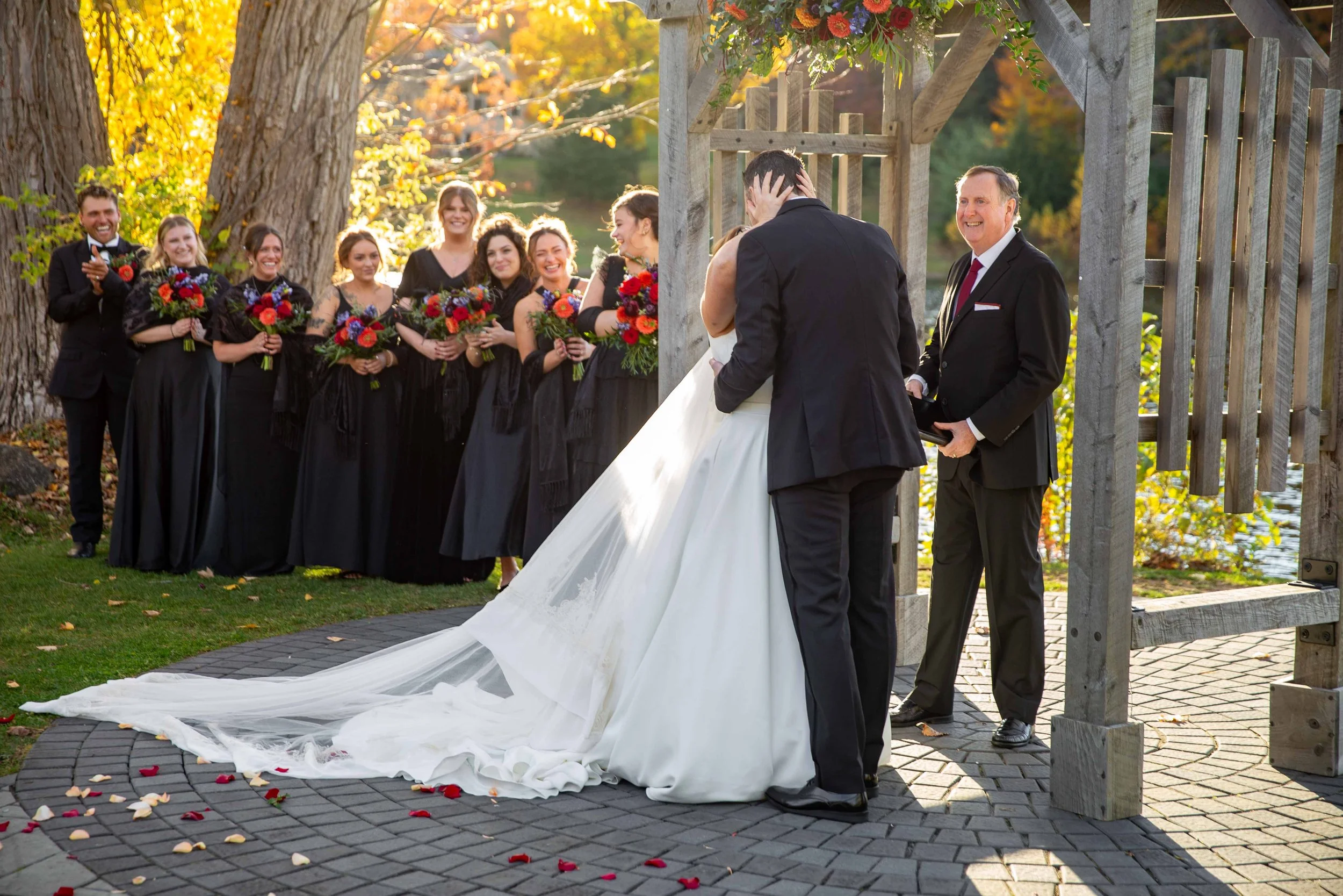 A wedding ceremony outdoors with a bride and groom exchanging vows under a wooden arch decorated with flowers, surrounded by bridesmaids in black dresses holding bouquets, and groomsmen applauding in the background, with autumn trees and sunlight cre