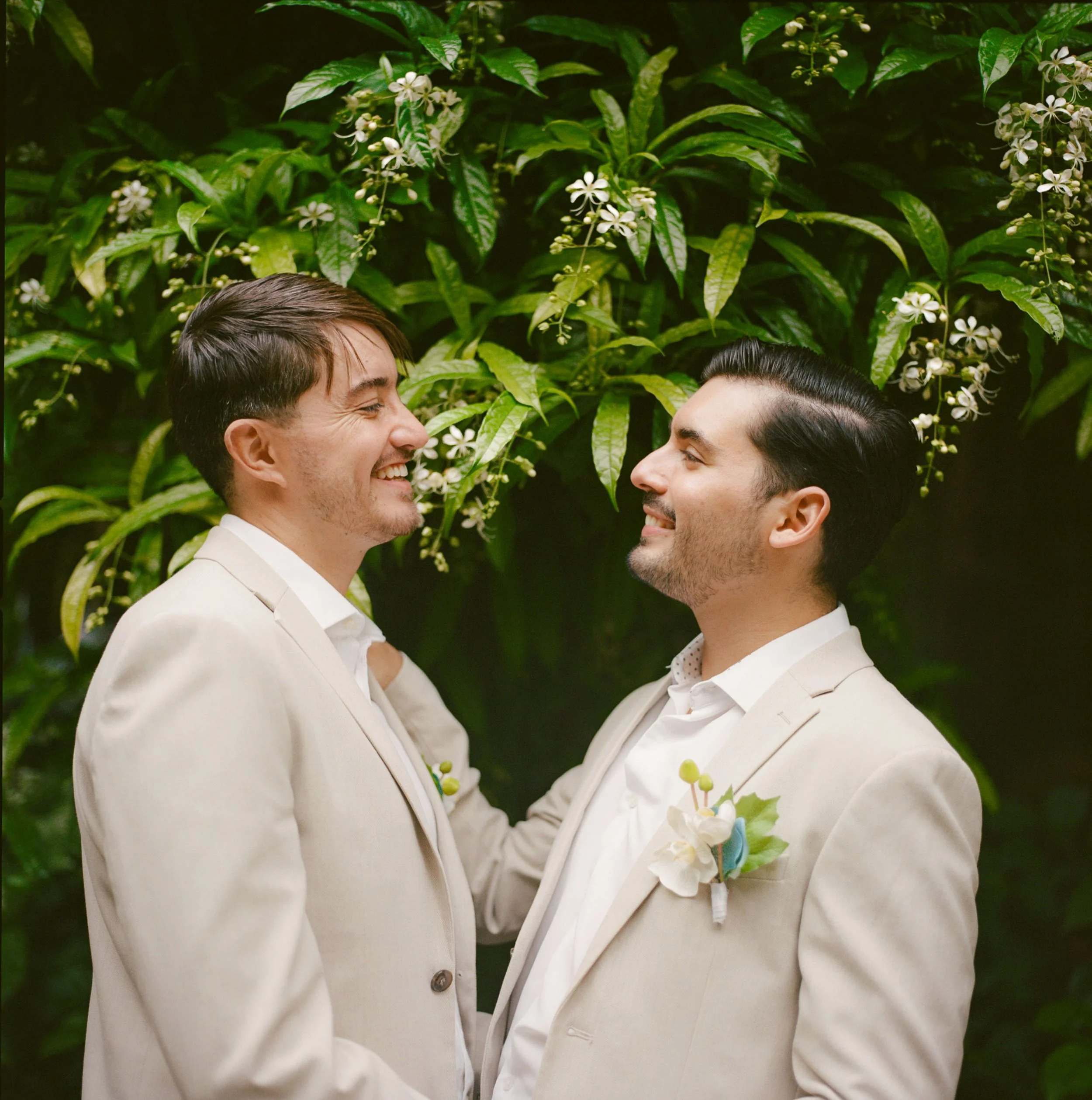 Two men in cream suits smiling at each other in front of green foliage and white flowers.