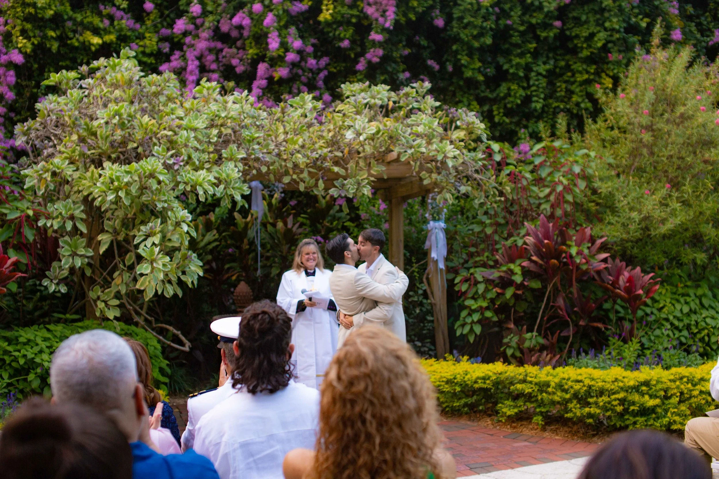 A same-sex couple is sharing a kiss during their outdoor wedding ceremony, with a band on a garden stage in the background and guests seated watching.