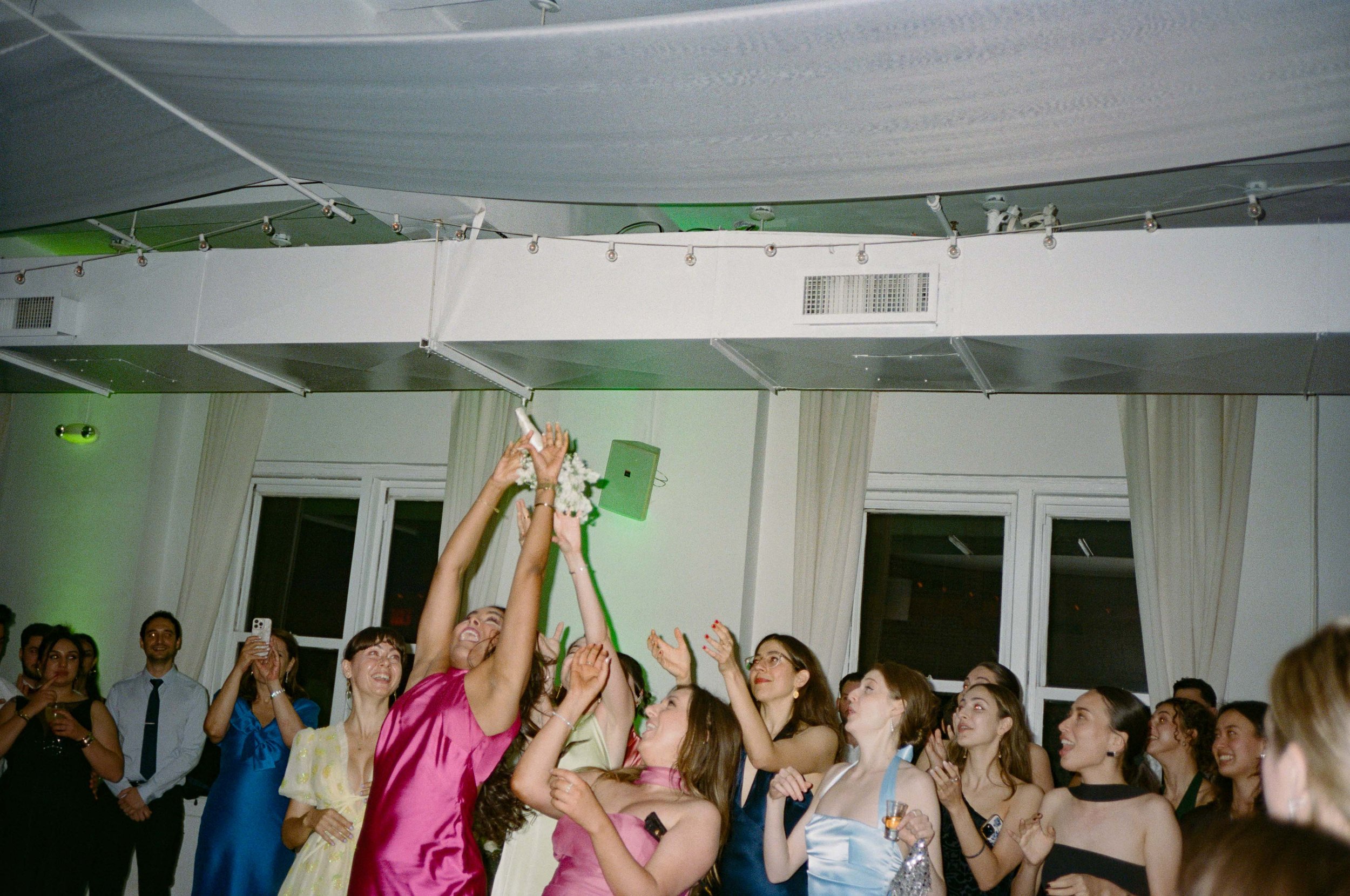 People at a celebration, some women in colorful dresses cheer as someone, wearing a bright pink dress, throws a bouquet in the air.