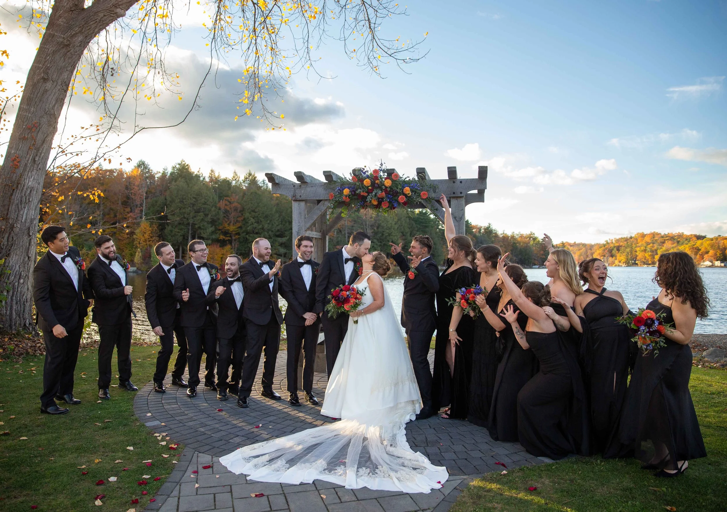A wedding ceremony outdoors by a lake during autumn, with the bride and groom kissing under a decorated wooden arch, surrounded by wedding party members dressed in black, celebrating and smiling.