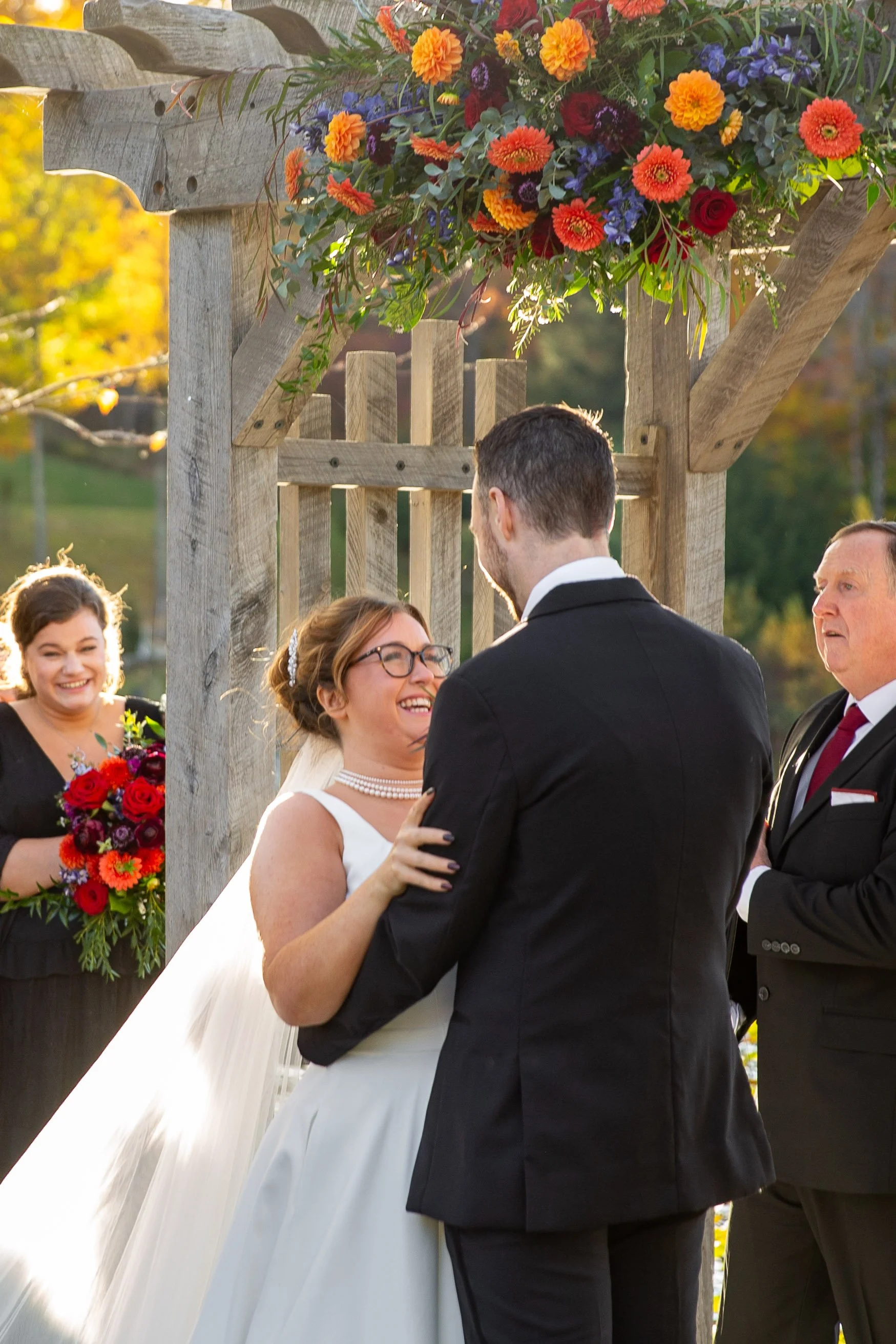 A bride and groom exchange vows at an outdoor wedding ceremony under a wooden arbor decorated with vibrant orange, red, and purple flowers, with a bridesmaid holding a bouquet in the background and a man officiating the ceremony.