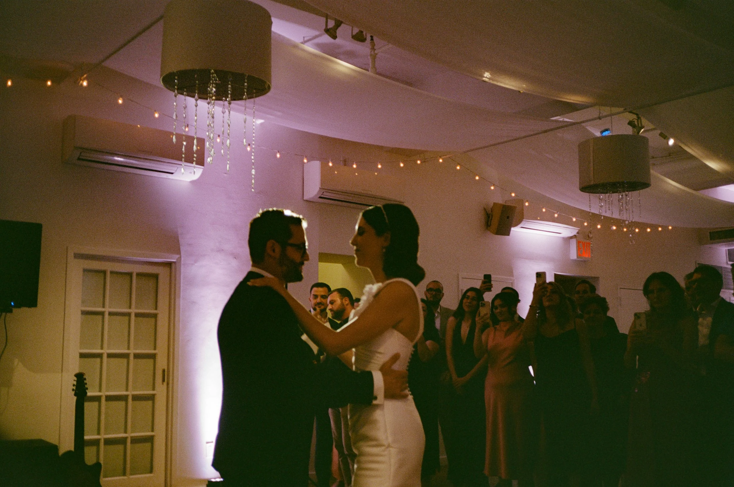 Couple dancing closely during their wedding reception with guests watching and taking photos.