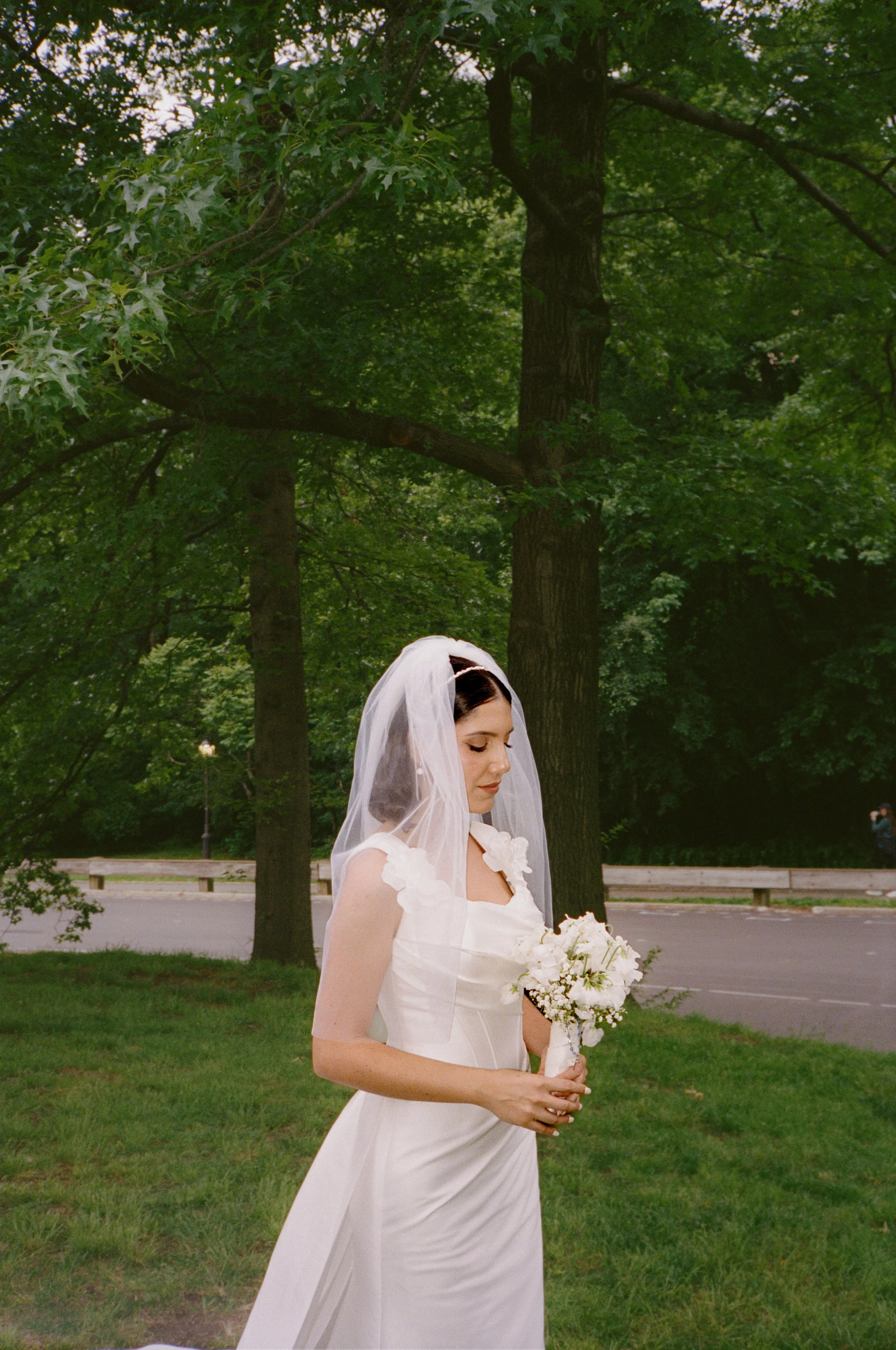 A bride in a white wedding dress with shoulder flower details, holding a bouquet of white flowers, and wearing a veil, standing outdoors near a large tree with green leaves.