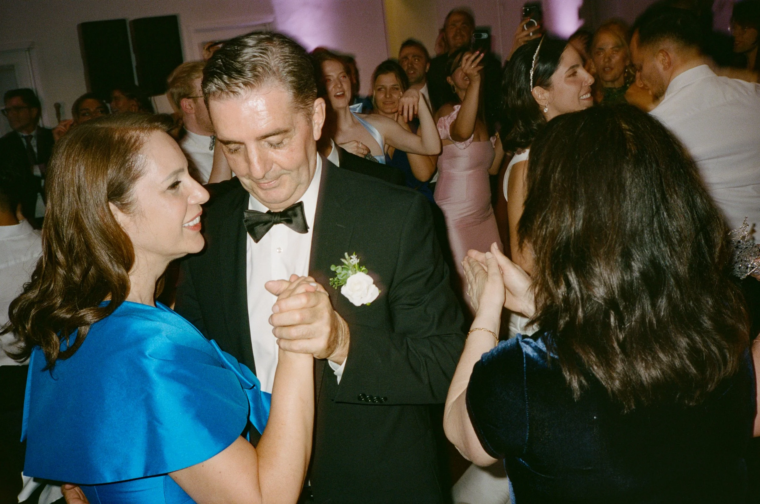 A couple dancing closely at a wedding reception, surrounded by other guests, some of whom are also dancing and taking photos.