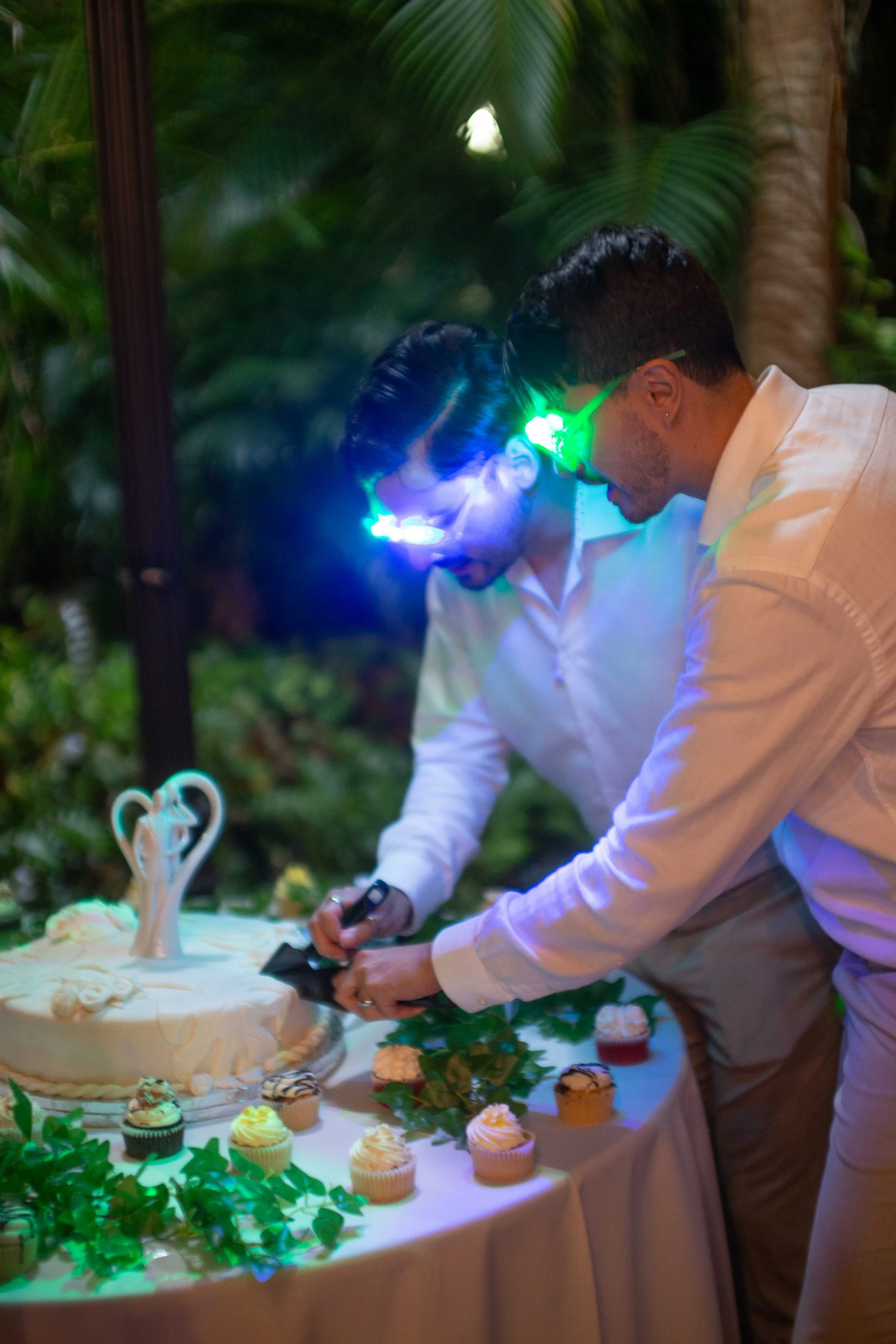 Two men in white shirts wearing glowing laser glasses, cutting a wedding cake at a celebration with cupcakes and green foliage on the table.