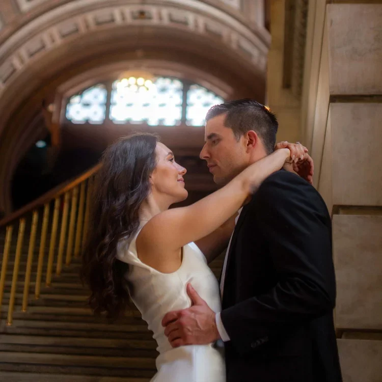 Engagement photos taken in the New York Public Library of a romantic couple dancing on a grand staircase in an elegant building with arched ceilings and a large window in the background.
