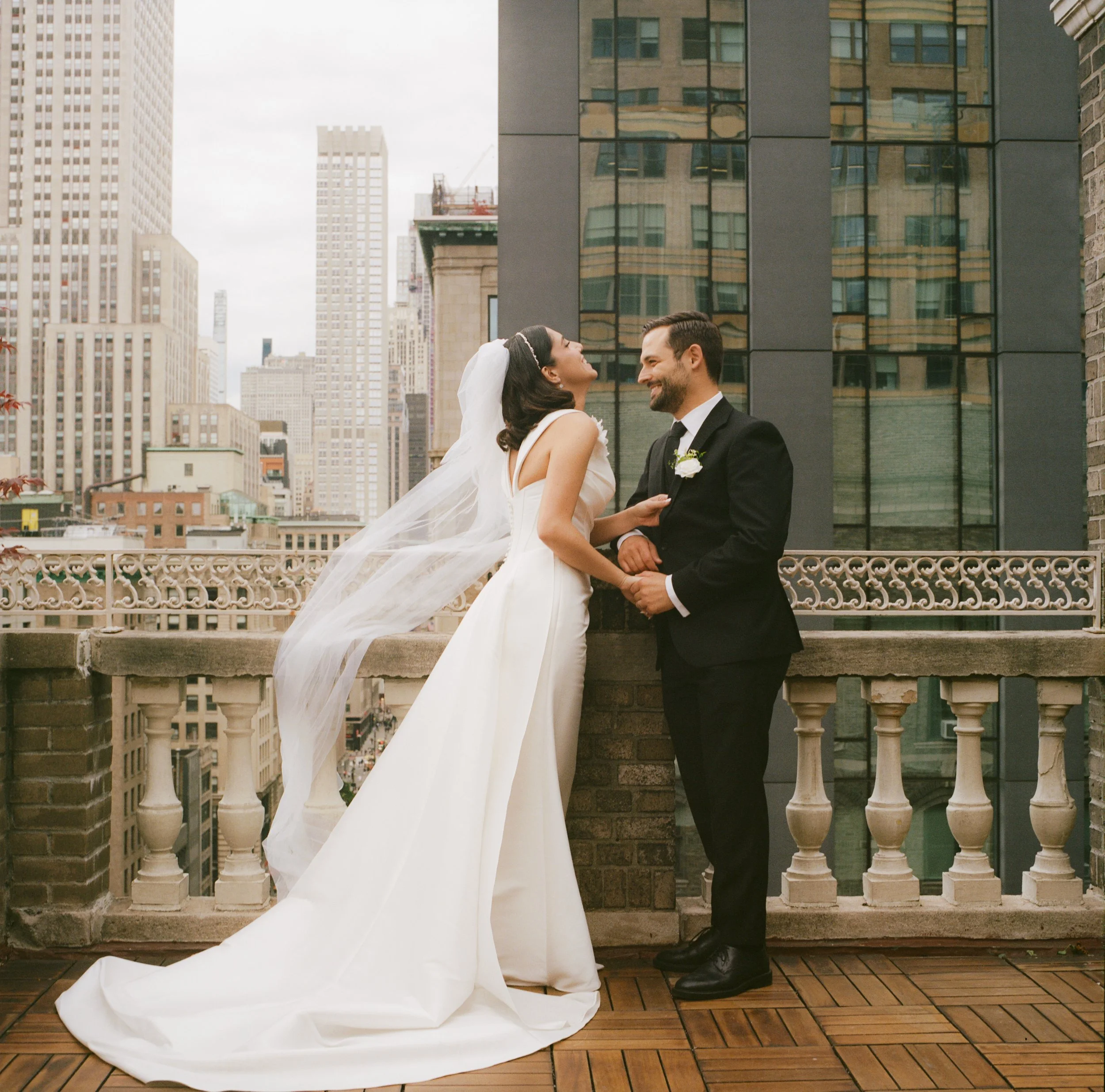 A bride and groom stand on a rooftop balcony overlooking a city, smiling at each other during their wedding photos.