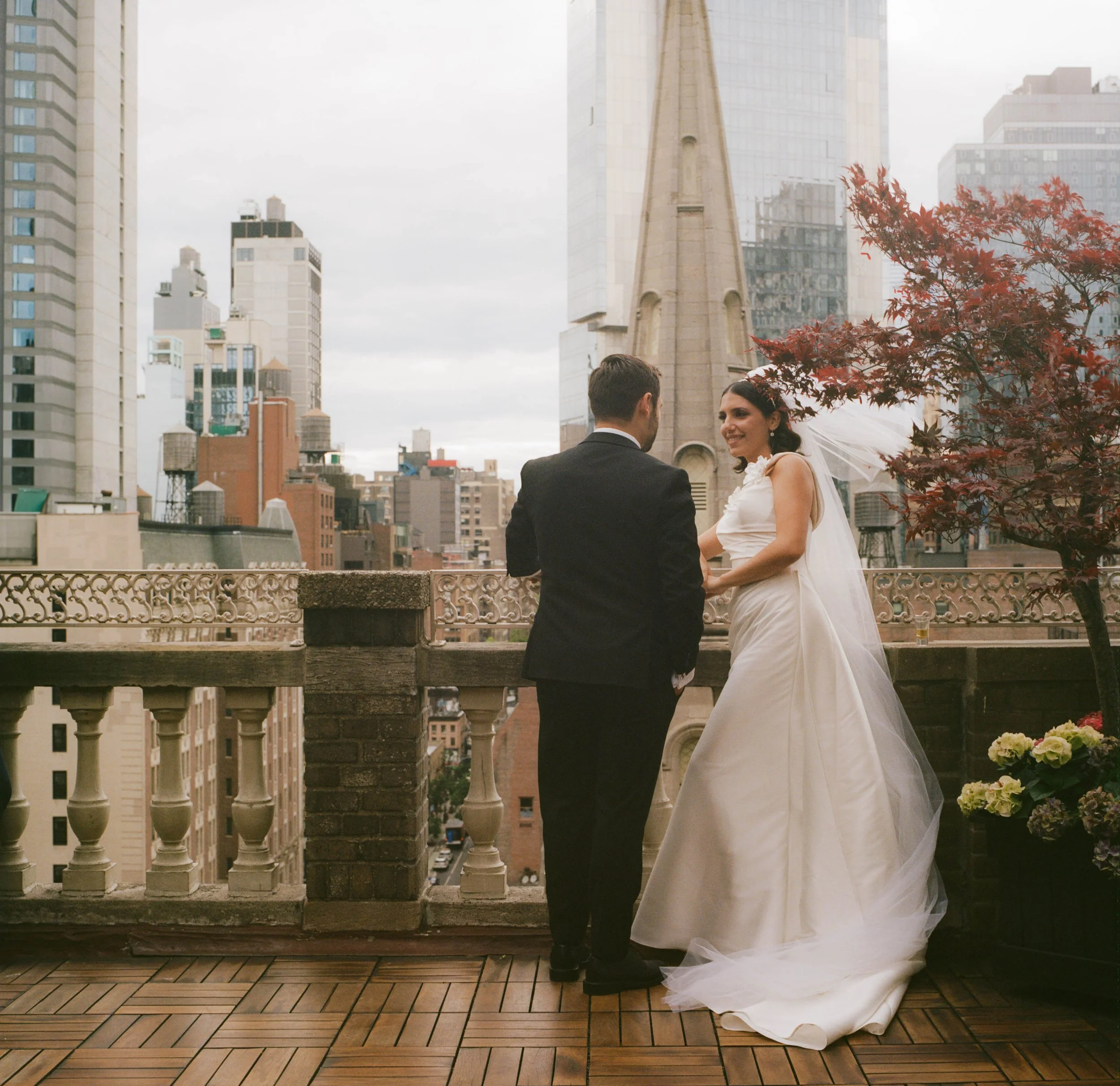 A bride and groom standing on a rooftop terrace with a city skyline in the background. The bride is wearing a white wedding gown and veil, and the groom is in a black suit. They are facing each other, holding hands, with a tree and flower pot nearby.
