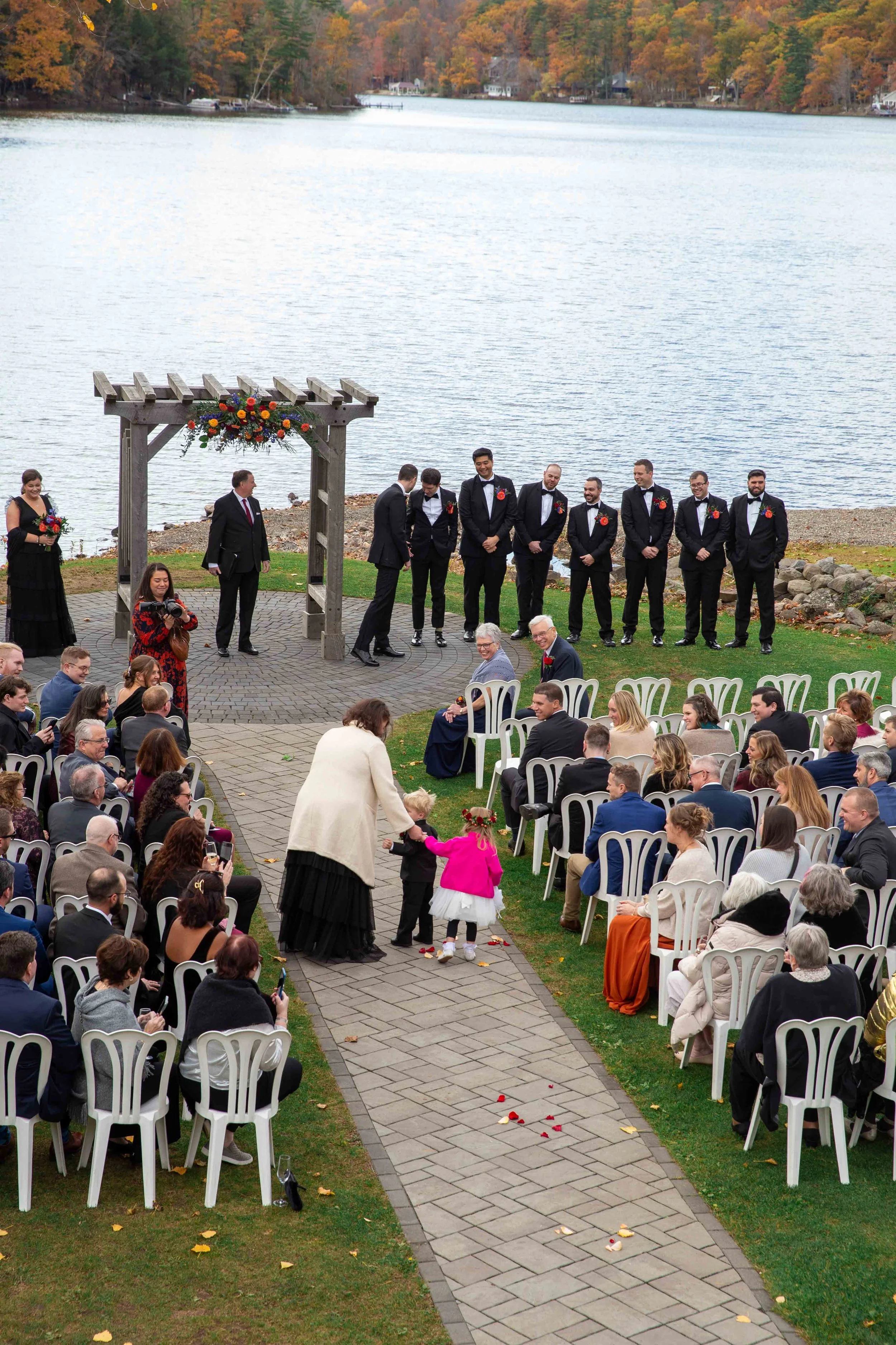 A lakeside outdoor wedding ceremony with guests seated, a couple at the altar, a wedding party in tuxedos, and a water and fall foliage background.