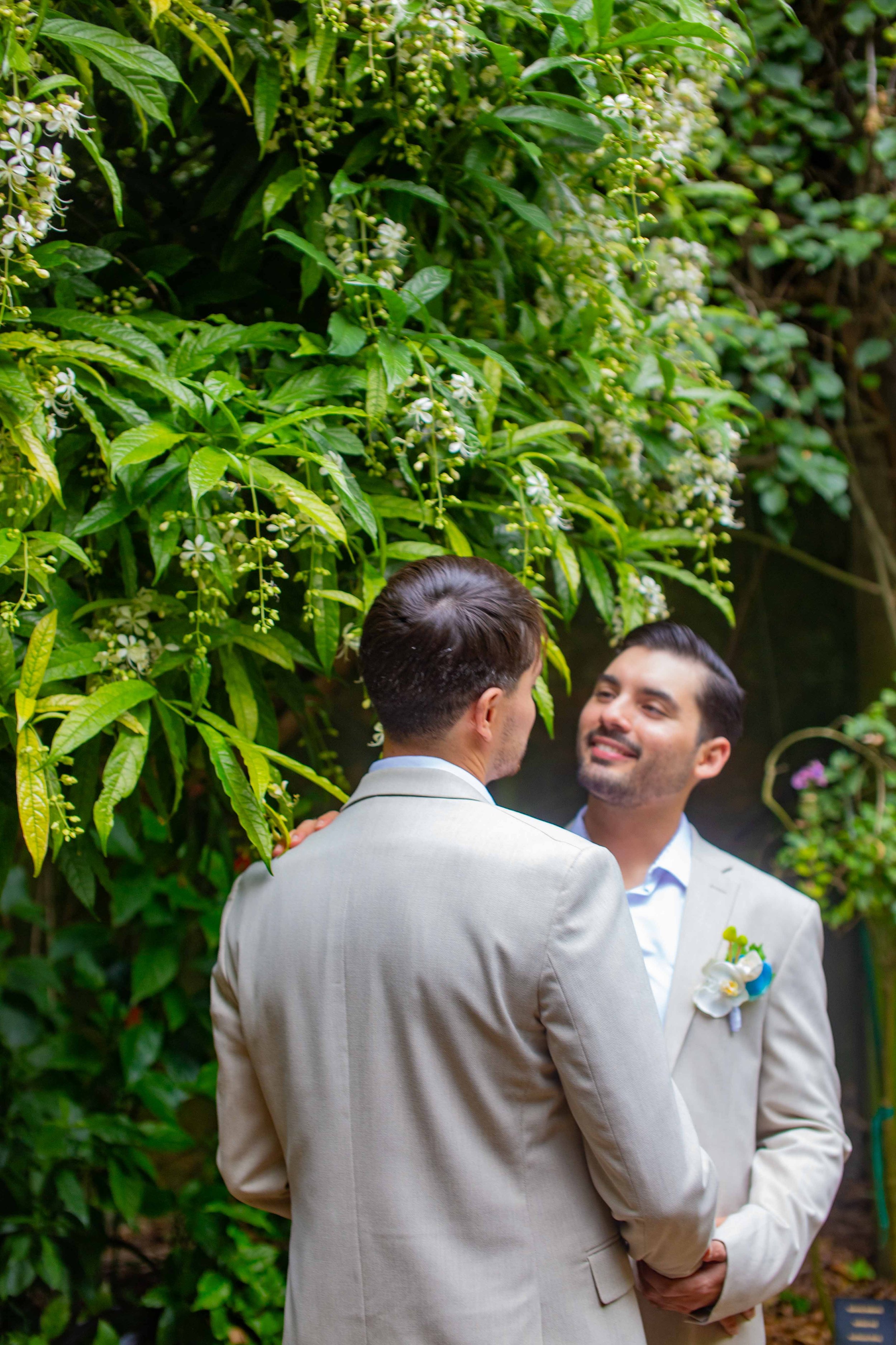 Two men wearing light-colored suits are standing face to face, smiling, with one of them touching the other’s shoulder, in front of a lush green background with white flowers.