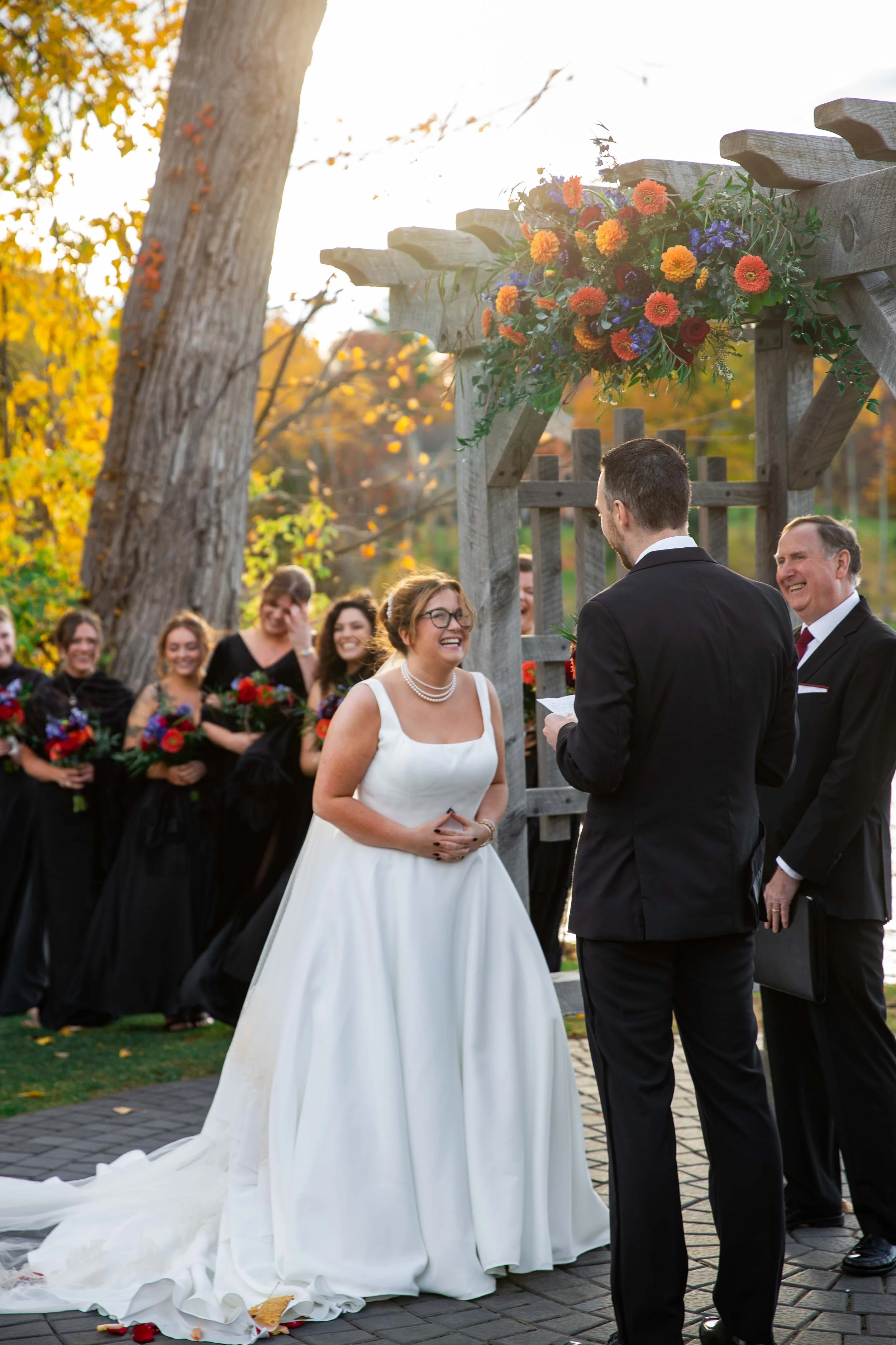 A bride and groom exchange vows during an outdoor wedding ceremony, with bridesmaids in black dresses holding bouquets in the background and officiant officiating, adorned with a floral arrangement overhead, in a scenic setting with fall foliage.