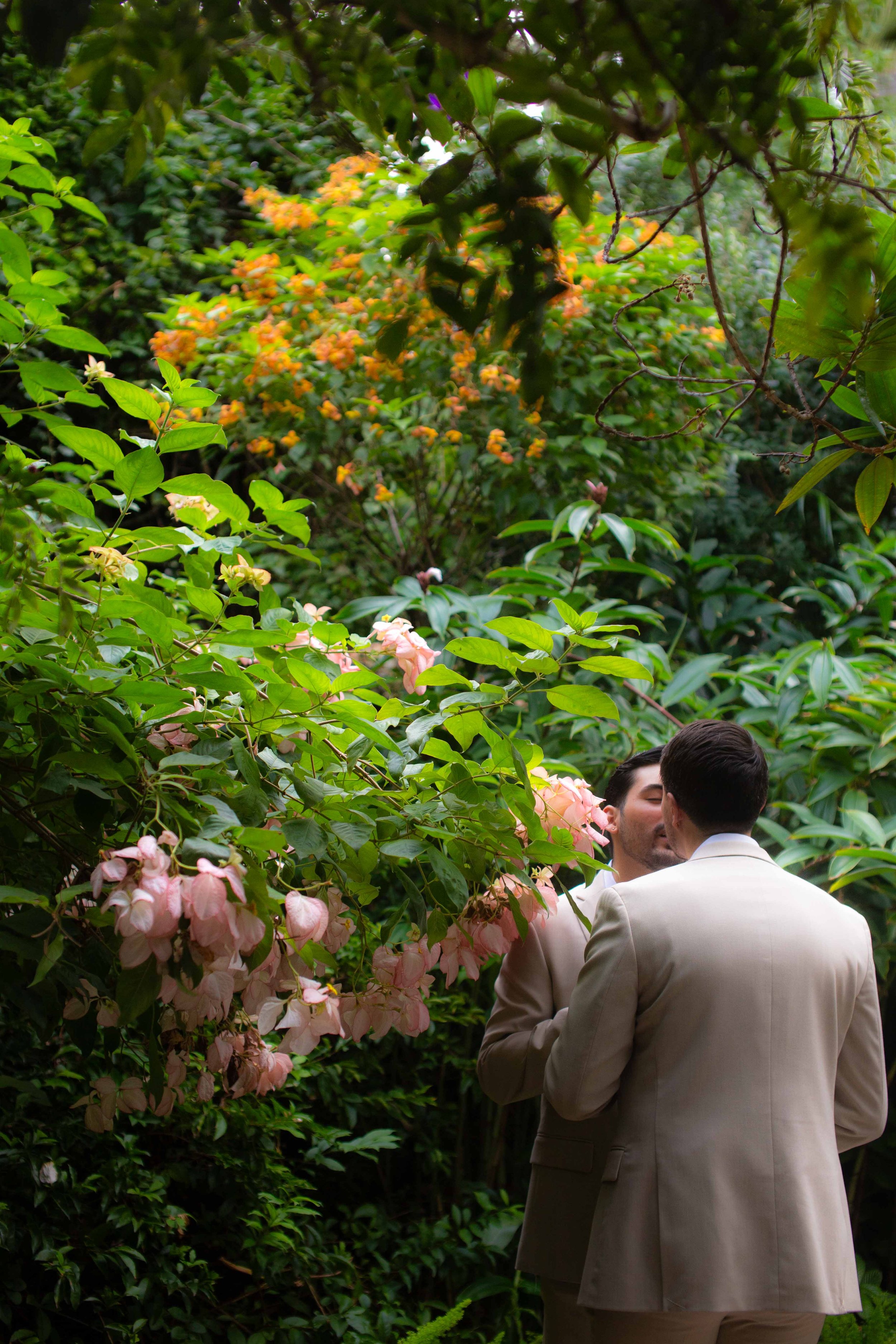 Two men in beige suits sharing a kiss in a lush green garden with pink and orange flowers.