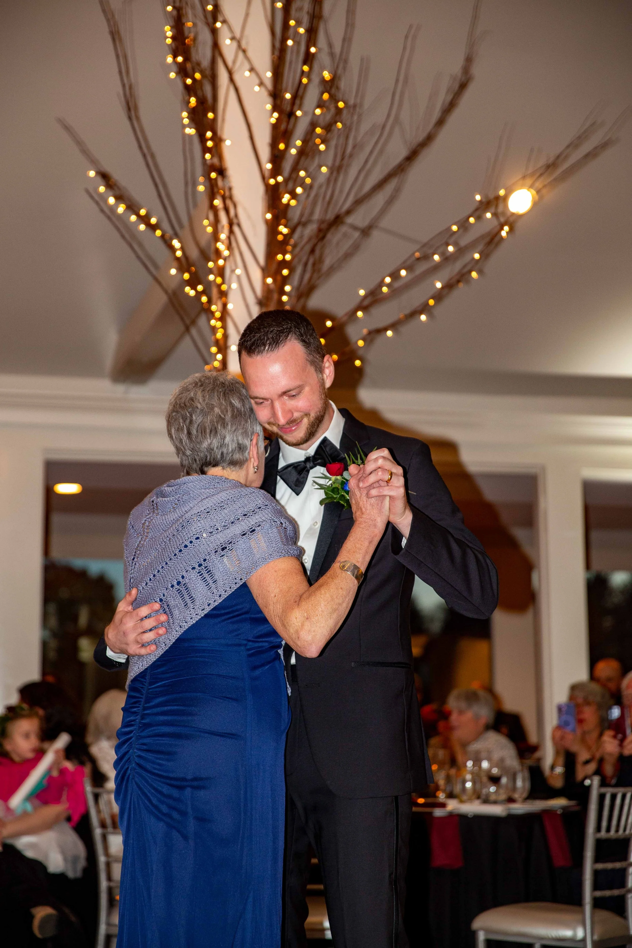 A young man in a tuxedo dancing with an older woman in a blue dress at a celebration or wedding reception, with guests seated at tables in the background under warm lighting.