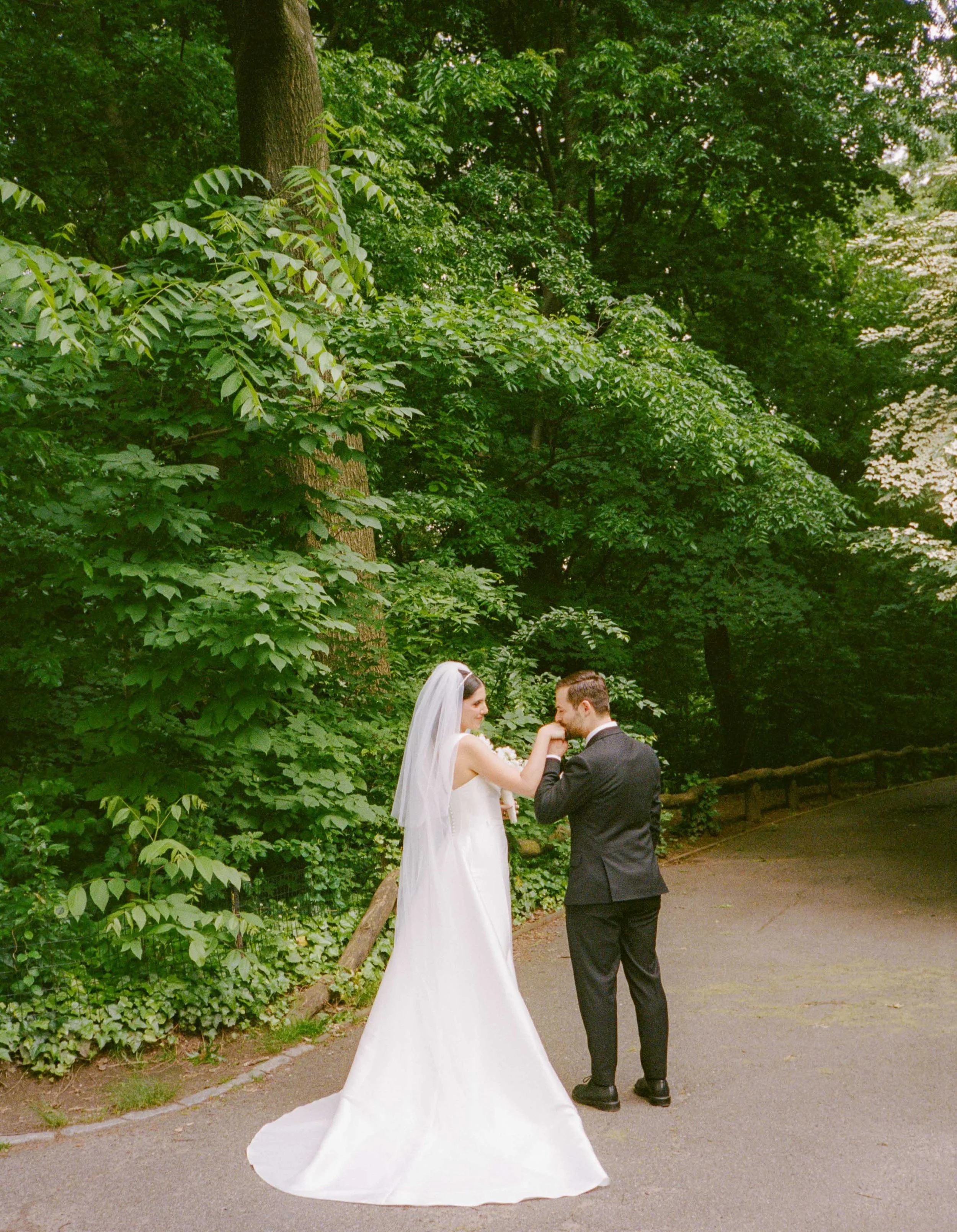A bride and groom holding hands on a path in a green, wooded park.