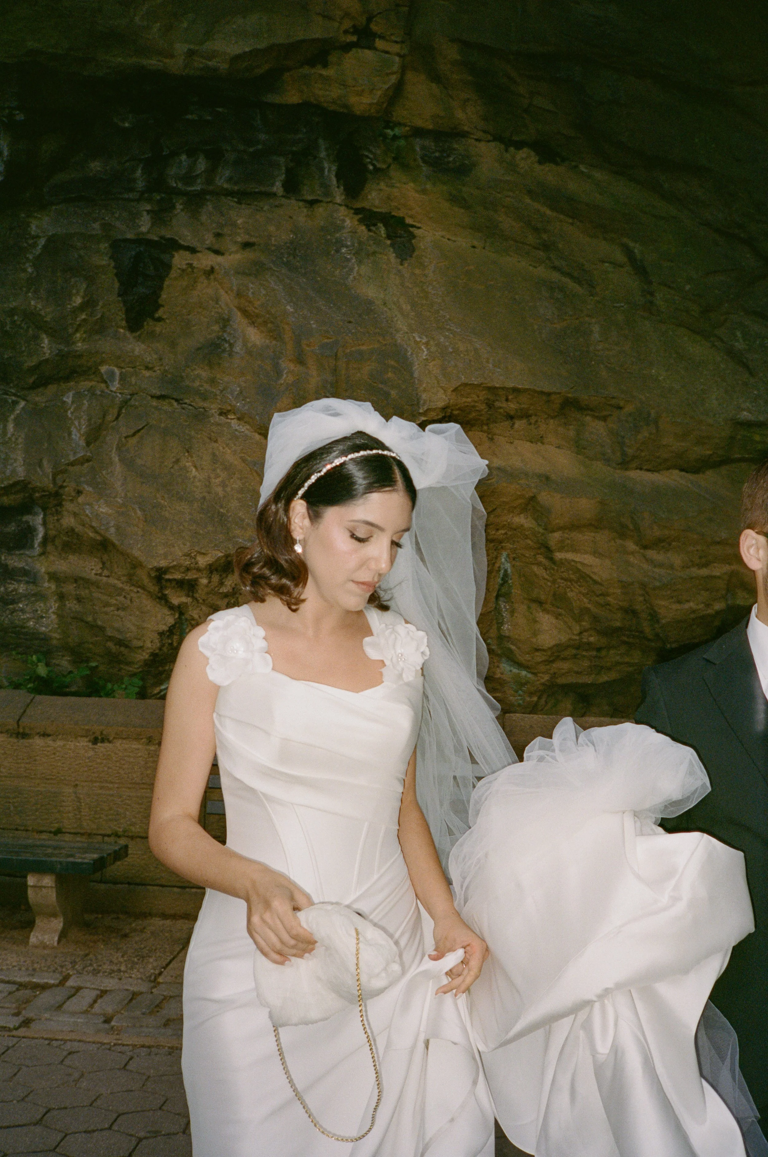 A bride in a white wedding dress holding a small white purse with a chain, standing next to a groom in a black tuxedo, in front of a rocky backdrop.