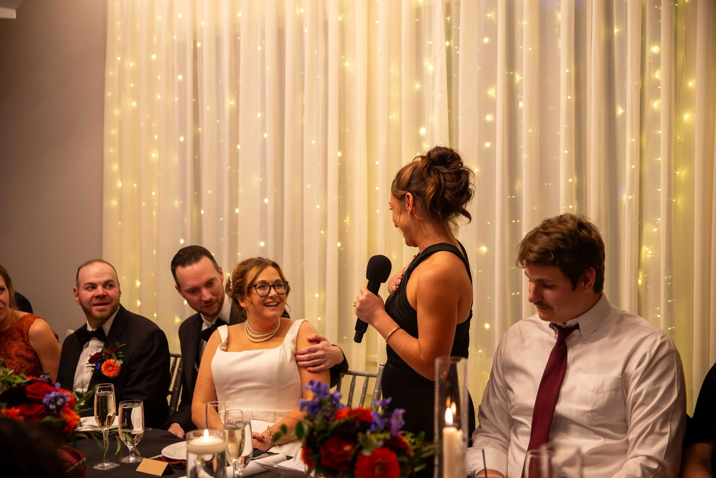 A woman in a black dress giving a speech at a wedding reception, with guests seated at a decorated table.