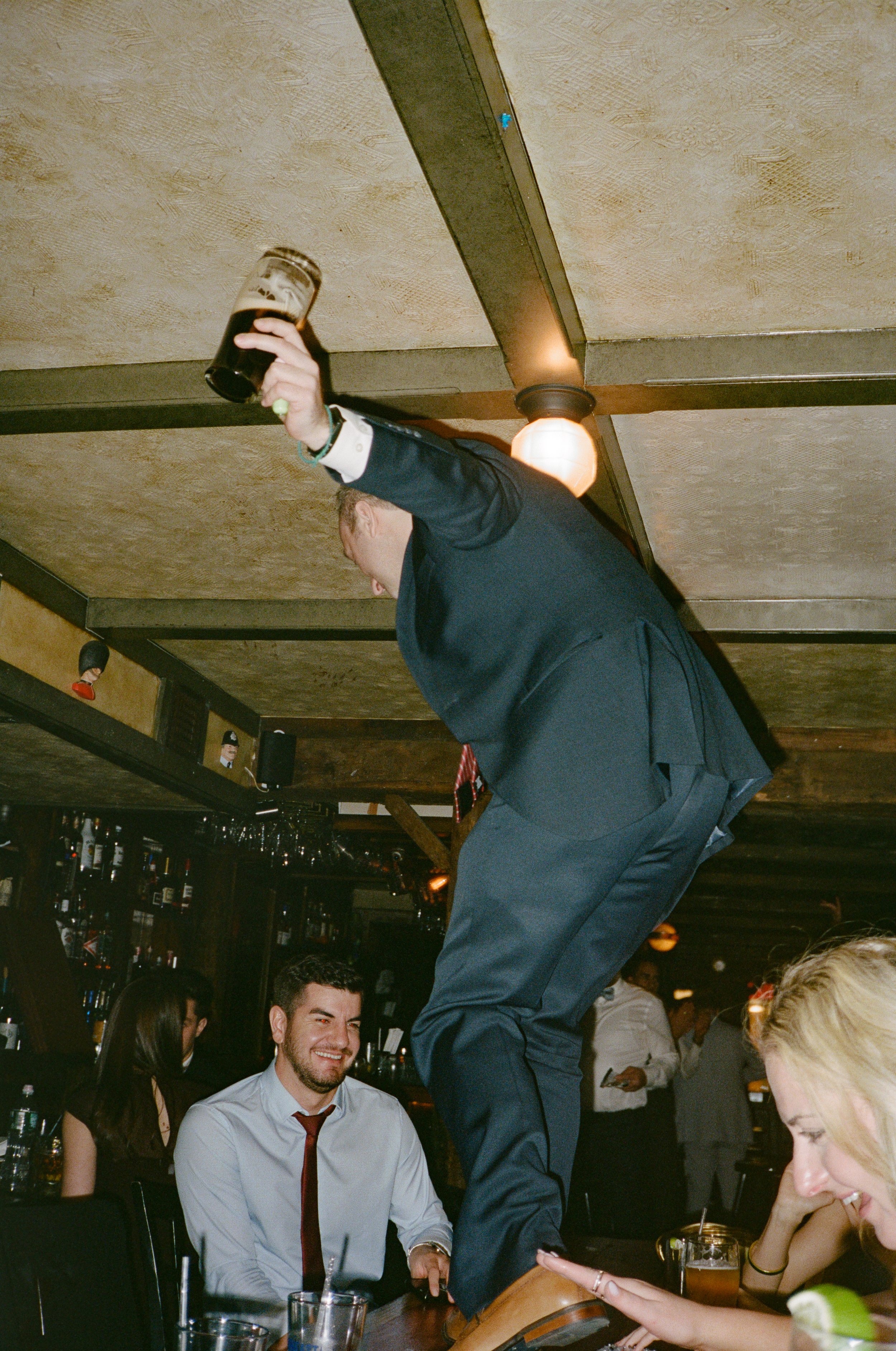 A man in a suit standing on a table in a bar, holding a beer, with other people sitting at the bar and smiling.