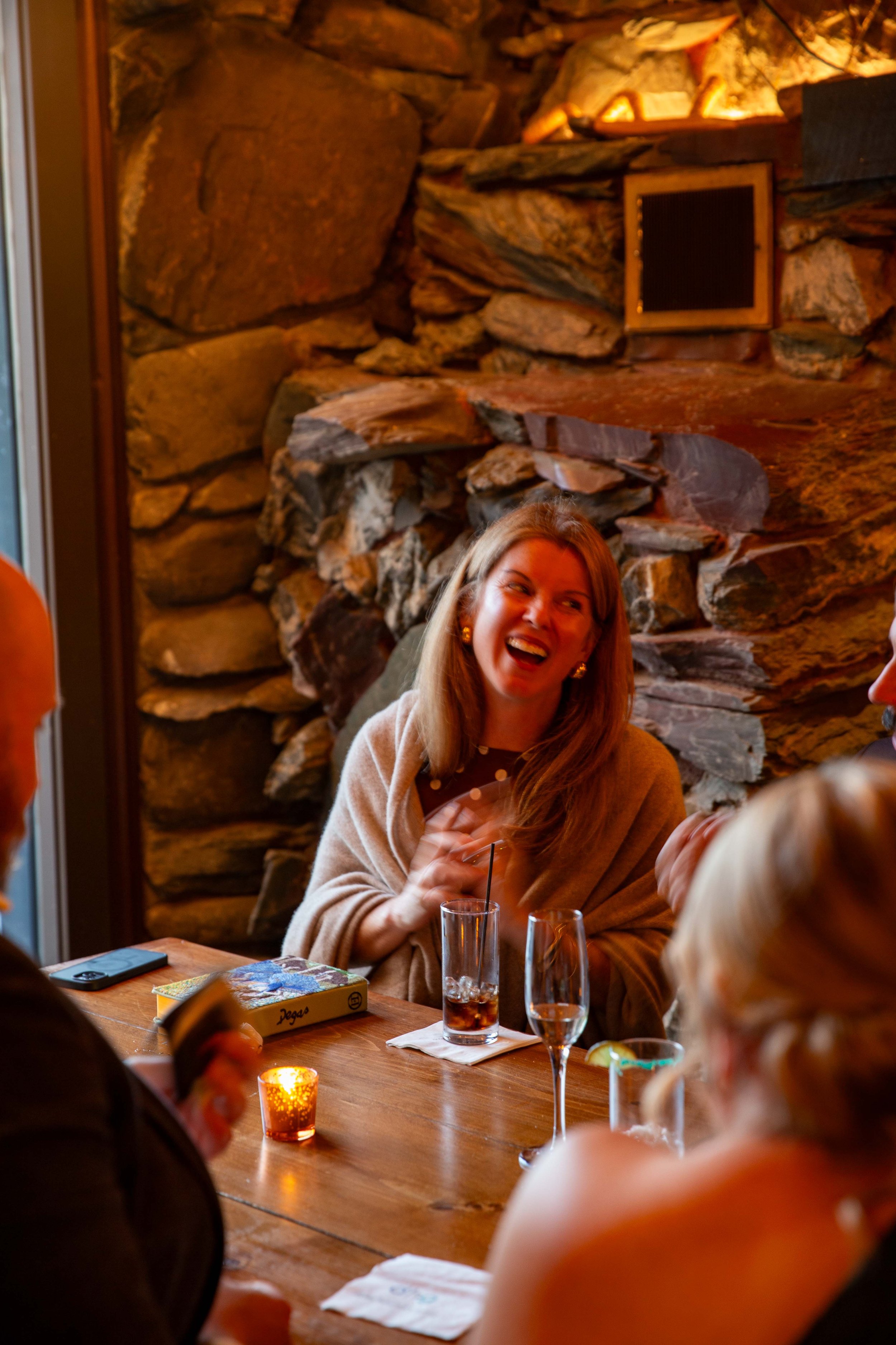 A woman laughing and enjoying drinks at a wooden table in a cozy, rustic restaurant with a stone fireplace in the background.