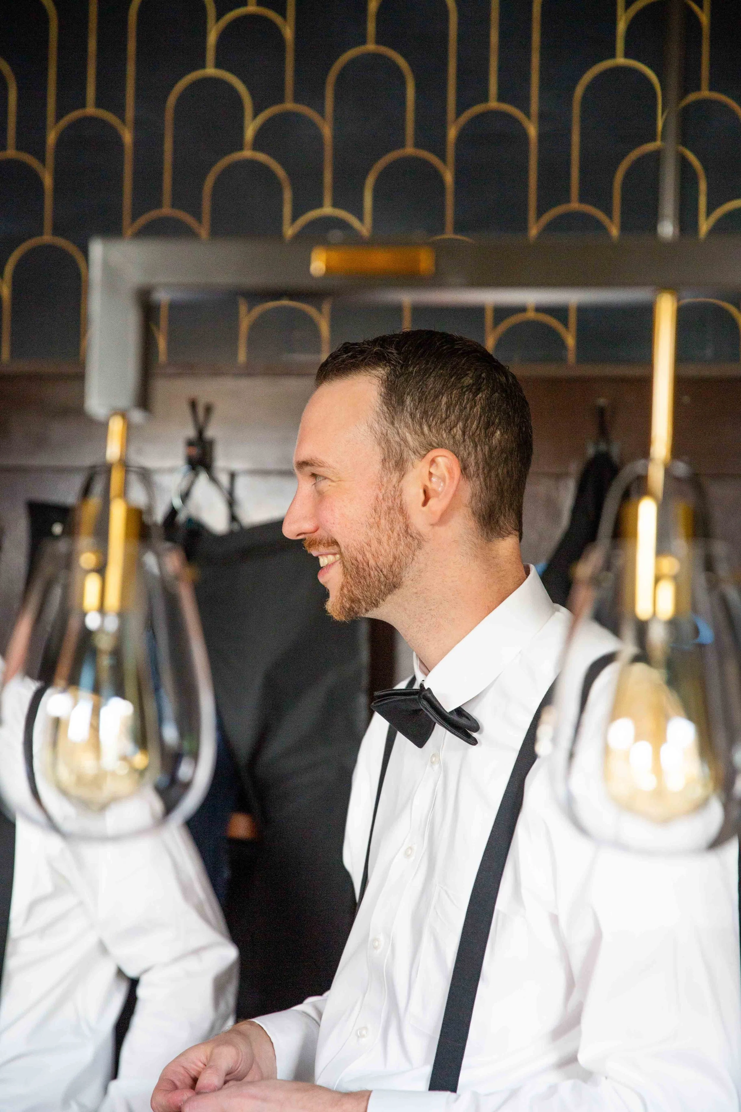 A smiling man dressed in formal attire, including a white shirt, black suspenders, and a black bow tie, is standing indoors near hanging light bulbs and a decorative wall.