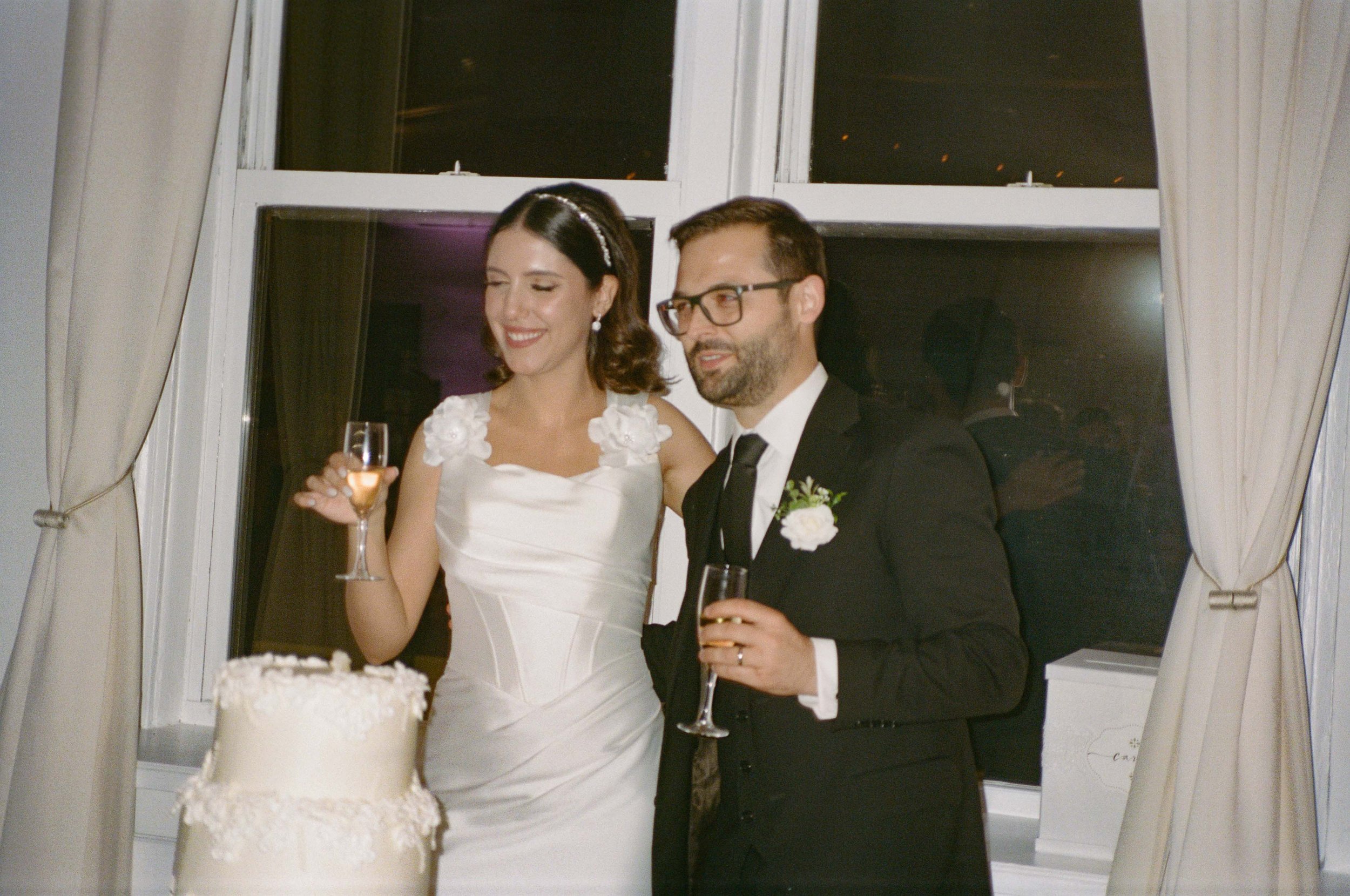 A bride and groom standing together at a wedding reception, holding champagne glasses, with a wedding cake in front of them. The bride is wearing a white dress with floral details on the shoulders, and the groom is in a black suit with a white shirt 