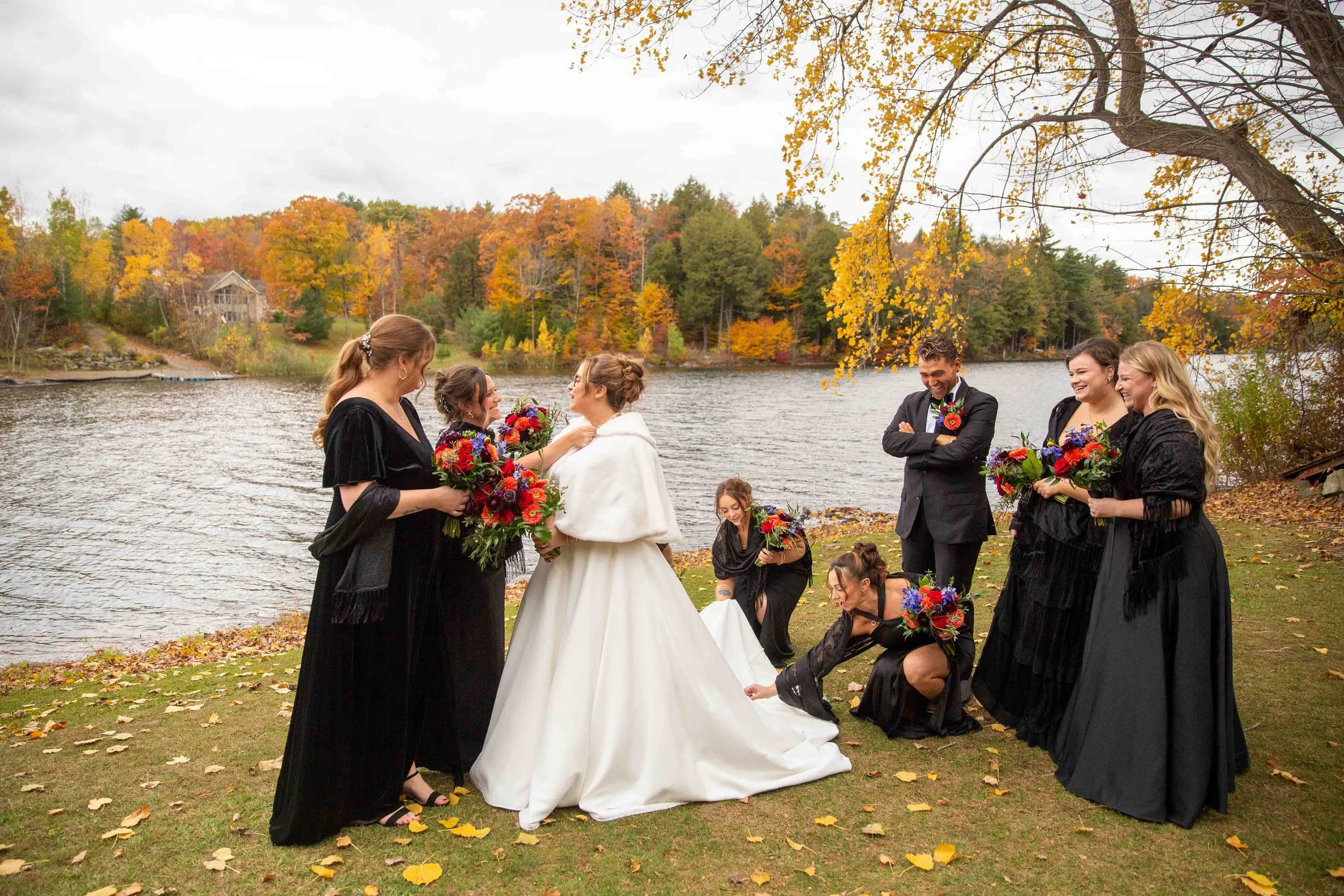 A wedding party by a lake with autumn trees. The bride is in a white gown, surrounded by bridesmaids in black dresses, holding colorful bouquets. Some women are adjusting the bride's dress, while men and women are smiling and laughing.