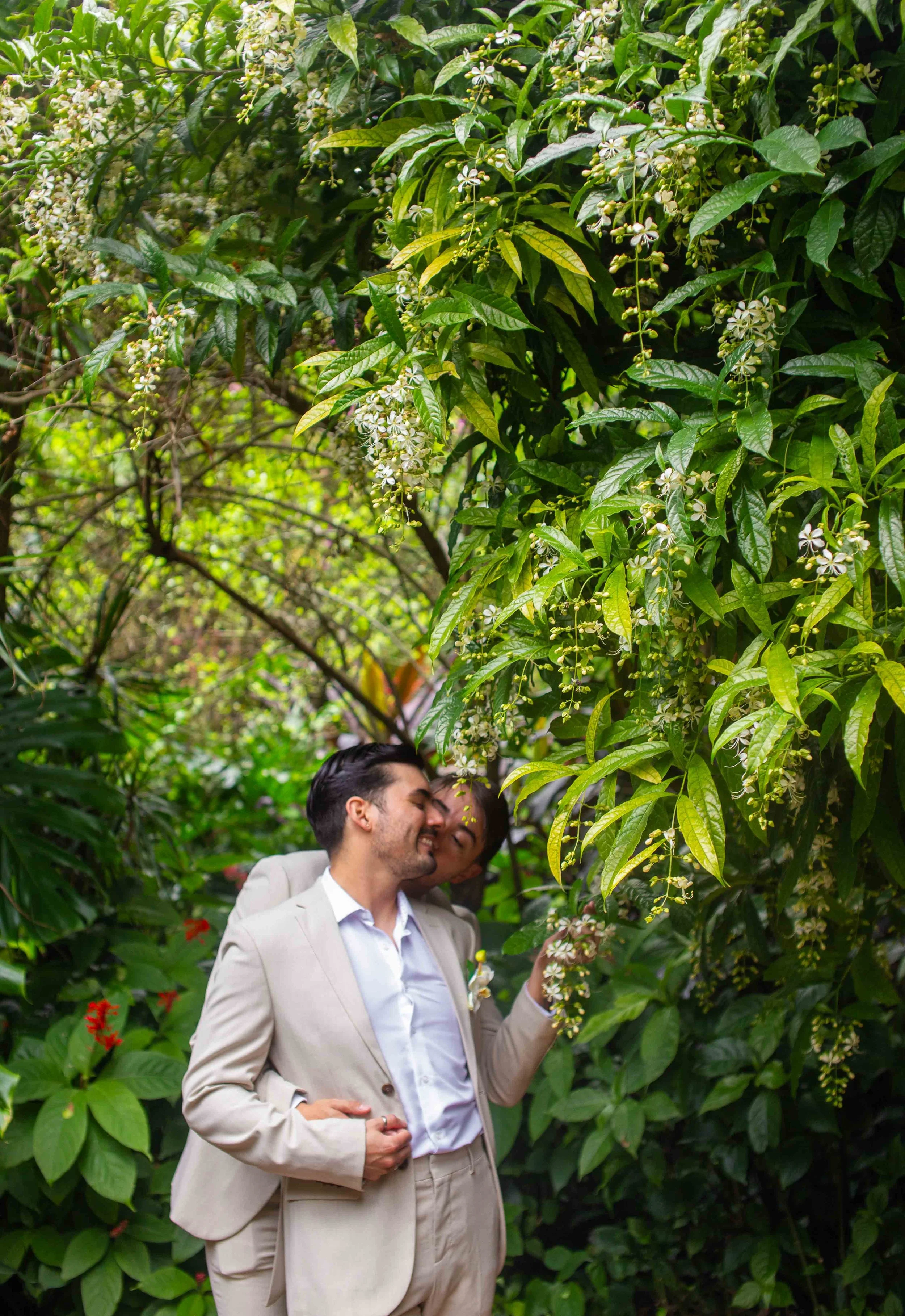 Two men in beige suits sharing a moment of happiness in a lush green garden surrounded by flowering plants.