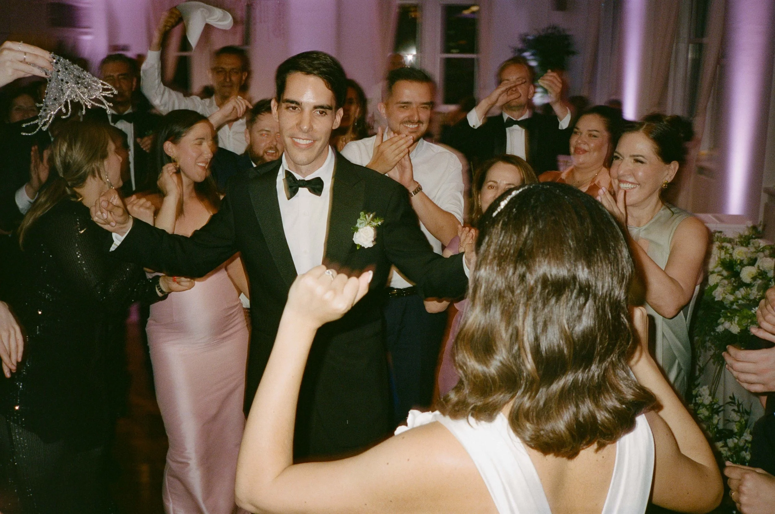 People dancing and celebrating at a wedding reception, with a groom in a tuxedo smiling in the center surrounded by friends and family.