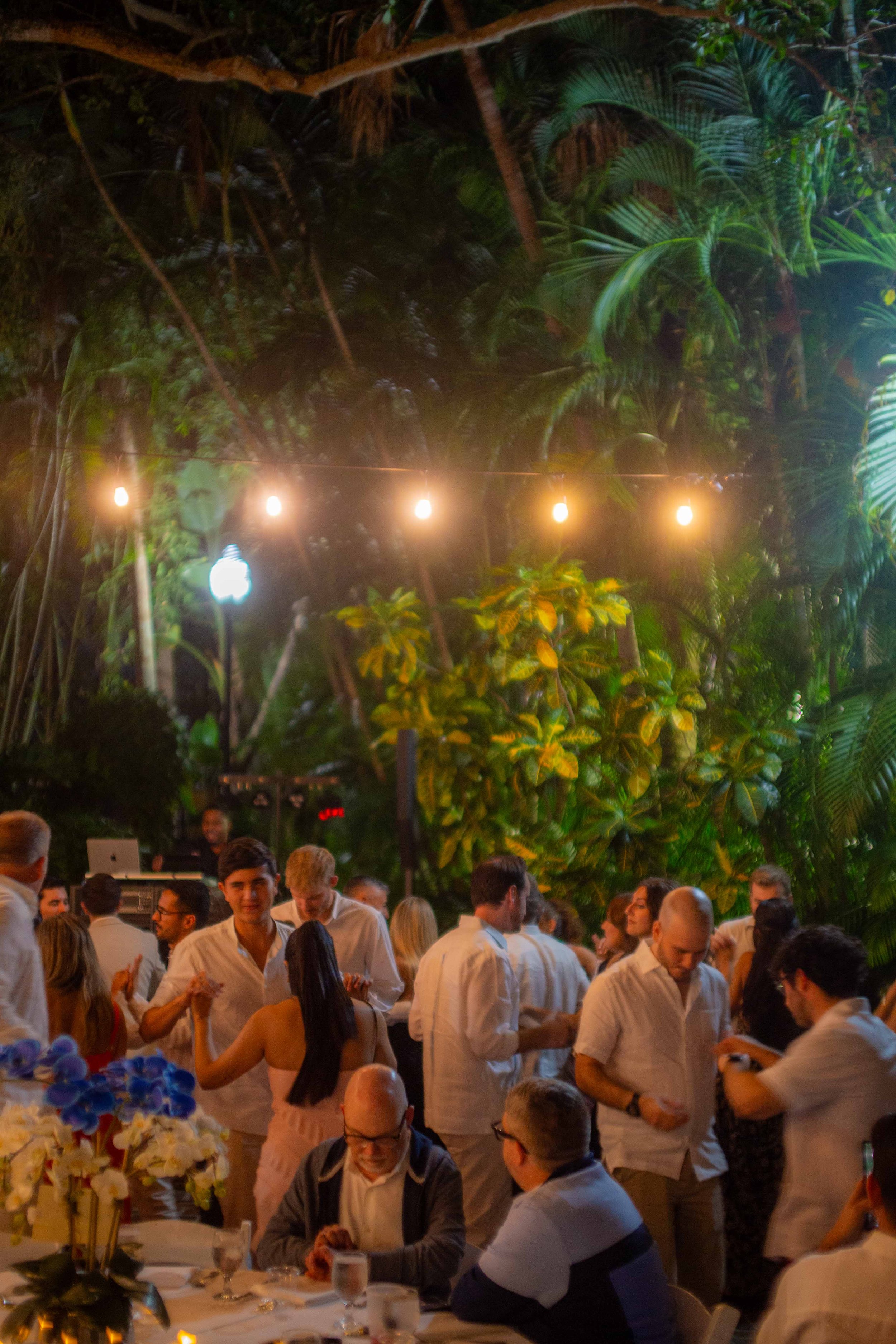 People dancing and socializing at an outdoor nighttime event surrounded by lush tropical plants, with string lights overhead.
