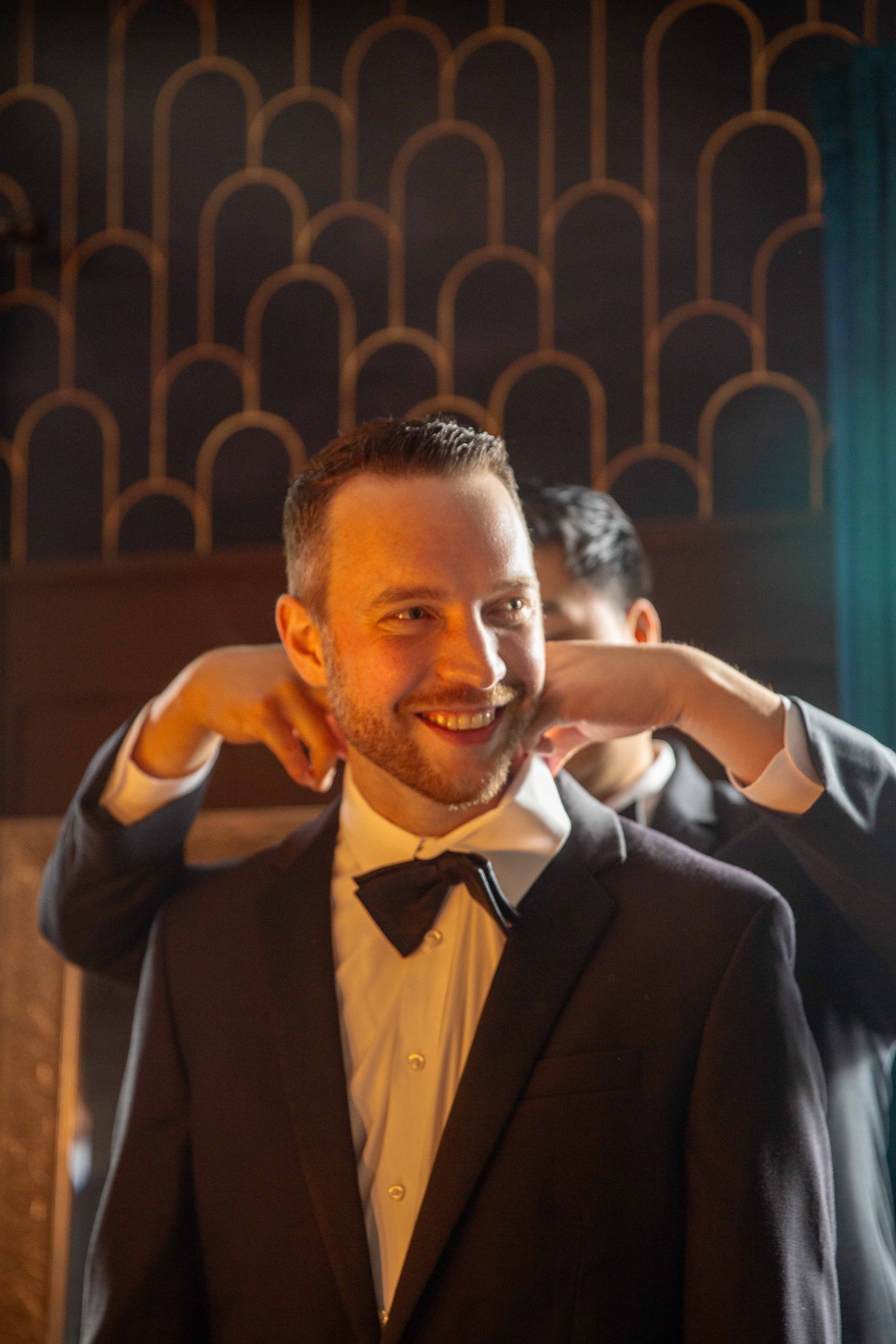 A man in formal attire with a tuxedo and bow tie, smiling in a warmly lit room, with another person in similar attire adjusting his collar.