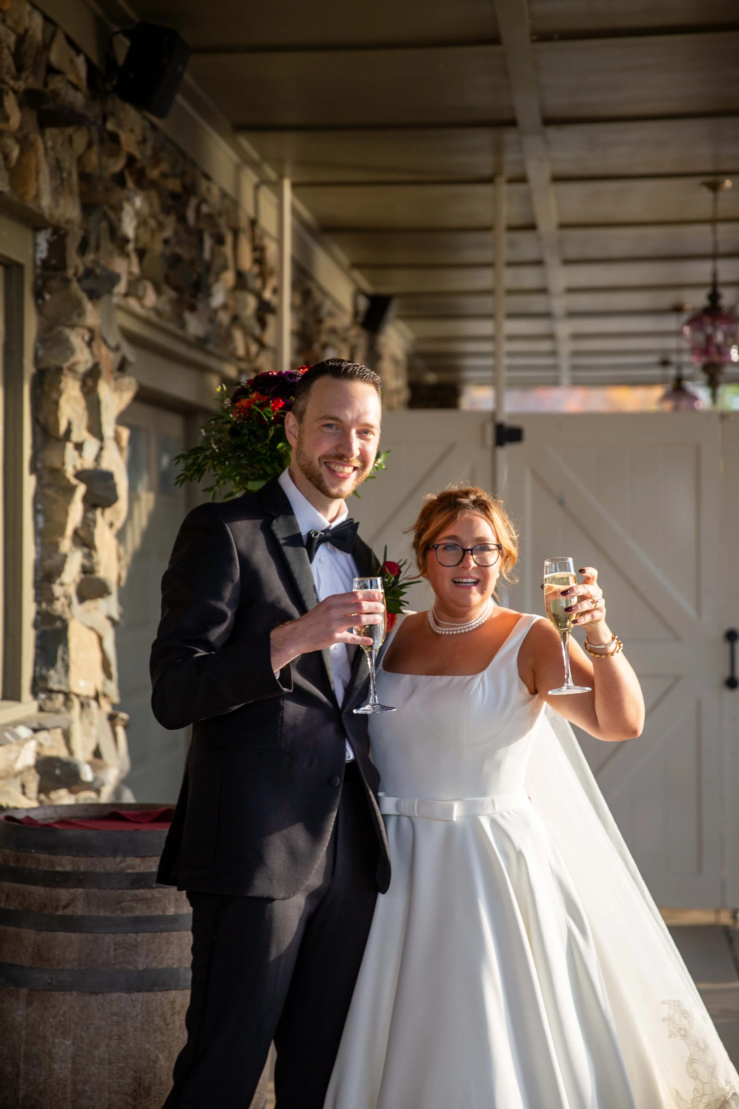A bride and groom celebrating with champagne during their wedding reception, smiling for the camera. The groom is wearing a black tuxedo with a bow tie, and the bride is in a white wedding dress with a pearl necklace and glasses, holding a glass of c