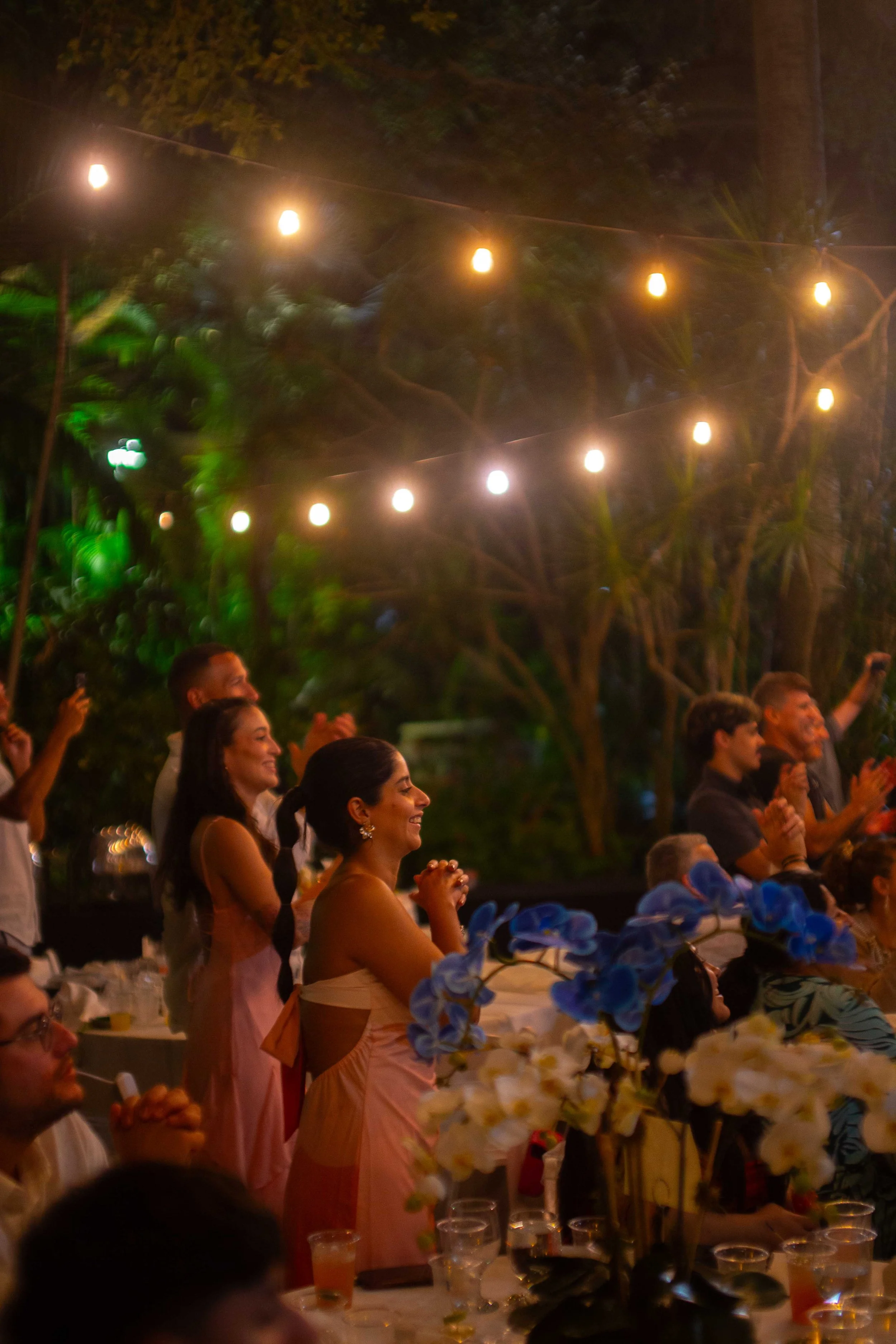 People at an outdoor celebration or event, smiling and clapping, with string lights overhead and floral arrangements on the table.