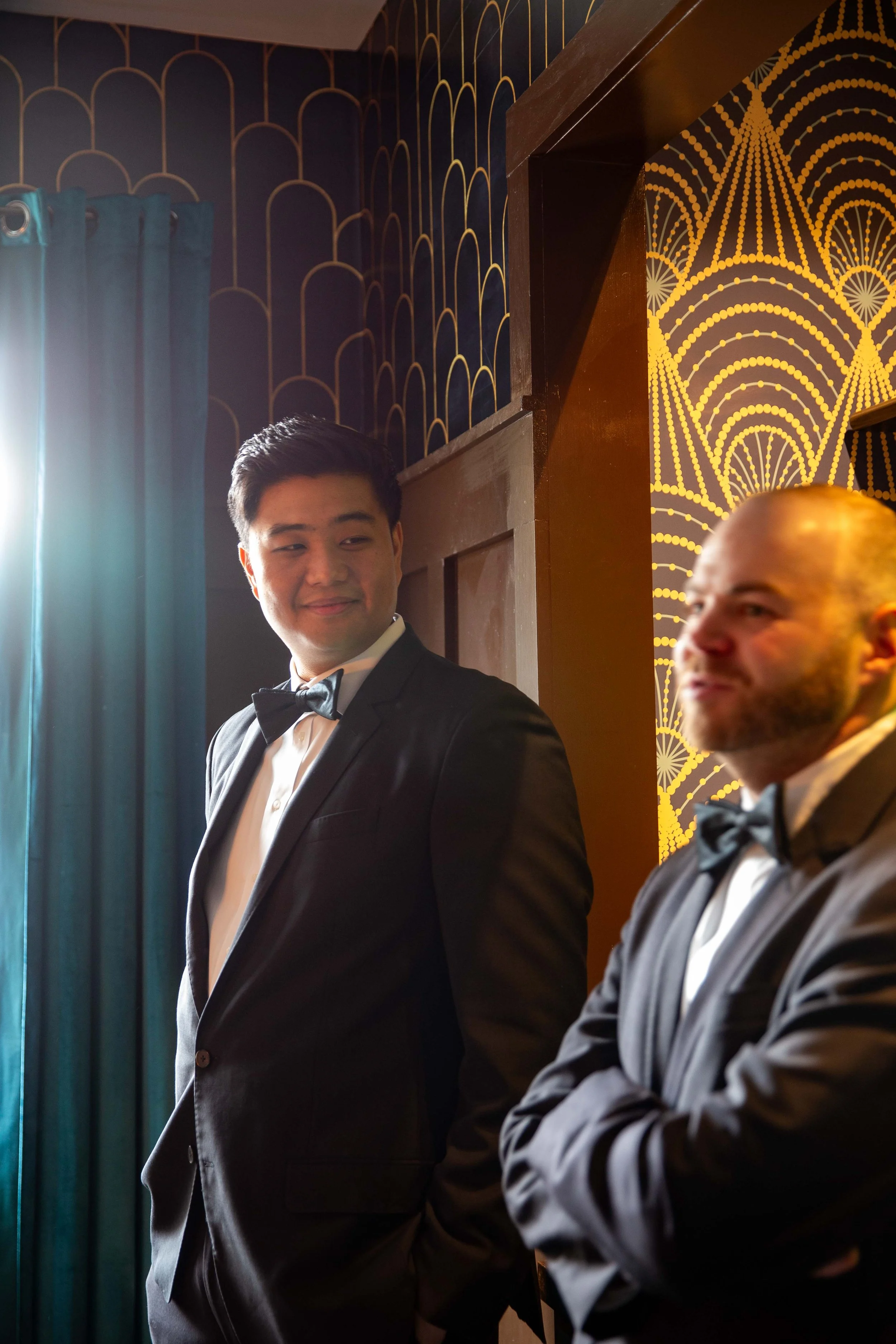 Two men in tuxedos with bow ties in an elegant room, one smiling and looking at the other, with dark walls decorated with gold patterns and a window with teal curtains.
