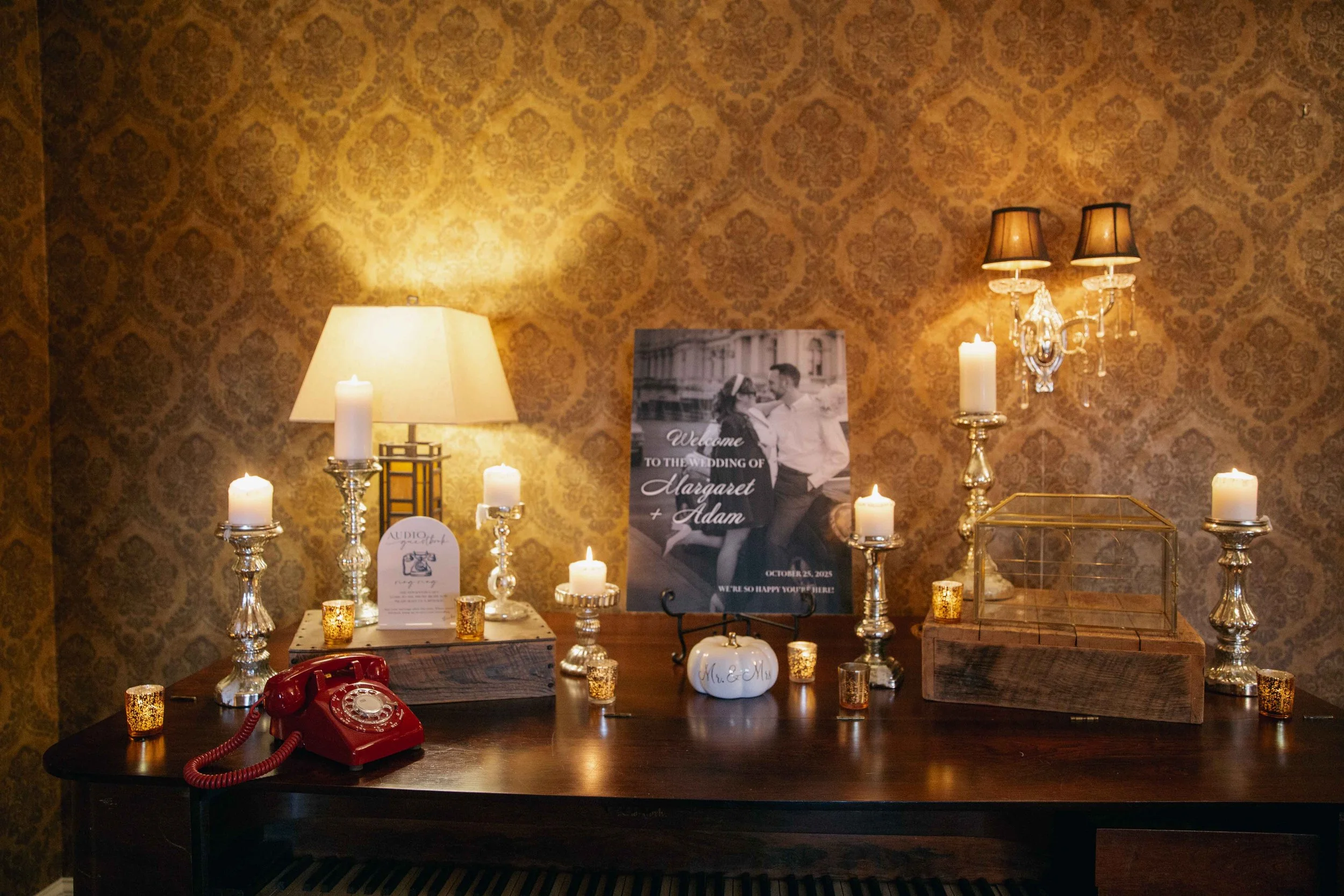 Wedding welcome display with candles, a vintage red rotary phone, a lamp, a framed sign with black and white photo of a couple, and a wooden box with a glass cover on a wooden table, against a gold patterned wall.