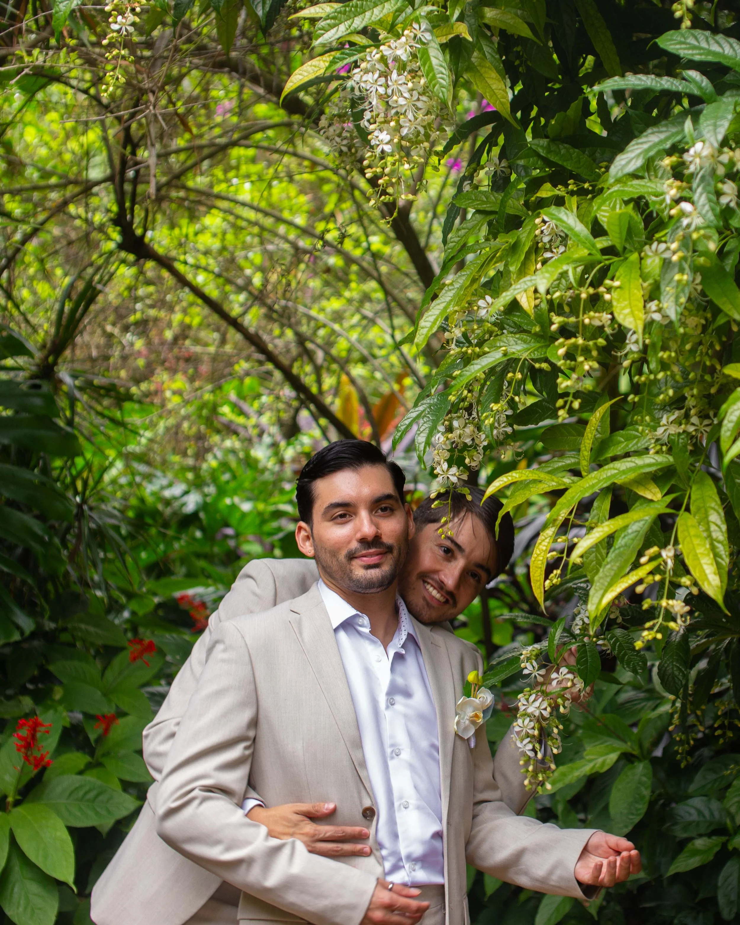 Two men in beige suits celebrating a wedding in a lush green garden, smiling and standing close together surrounded by tropical plants and flowers.