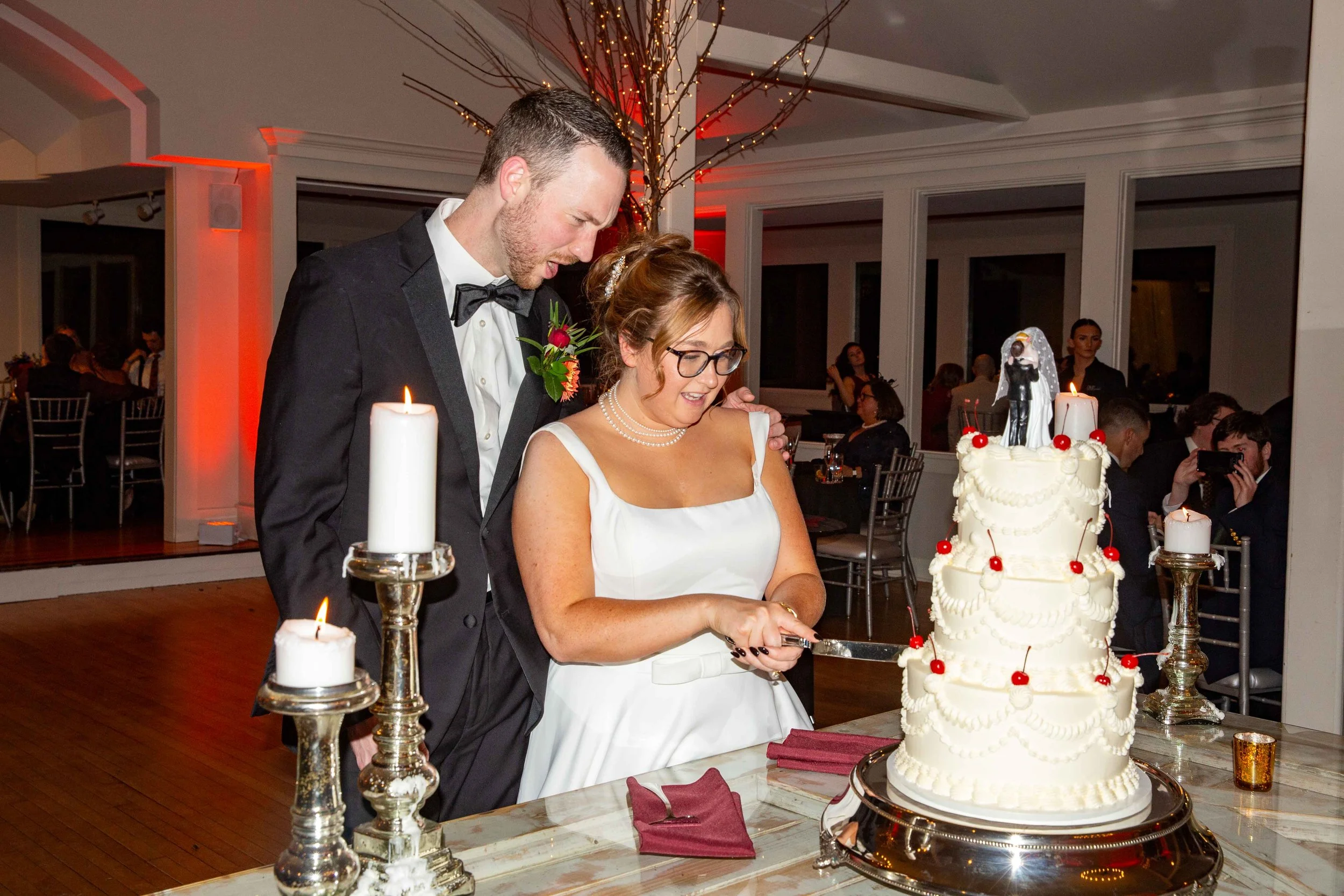 A bride and groom cutting a wedding cake together at their reception. The bride is wearing glasses and a white dress, while the groom is in a tuxedo with a boutonniere. The wedding cake is tall, white with red cherries and a bride and groom topper. C