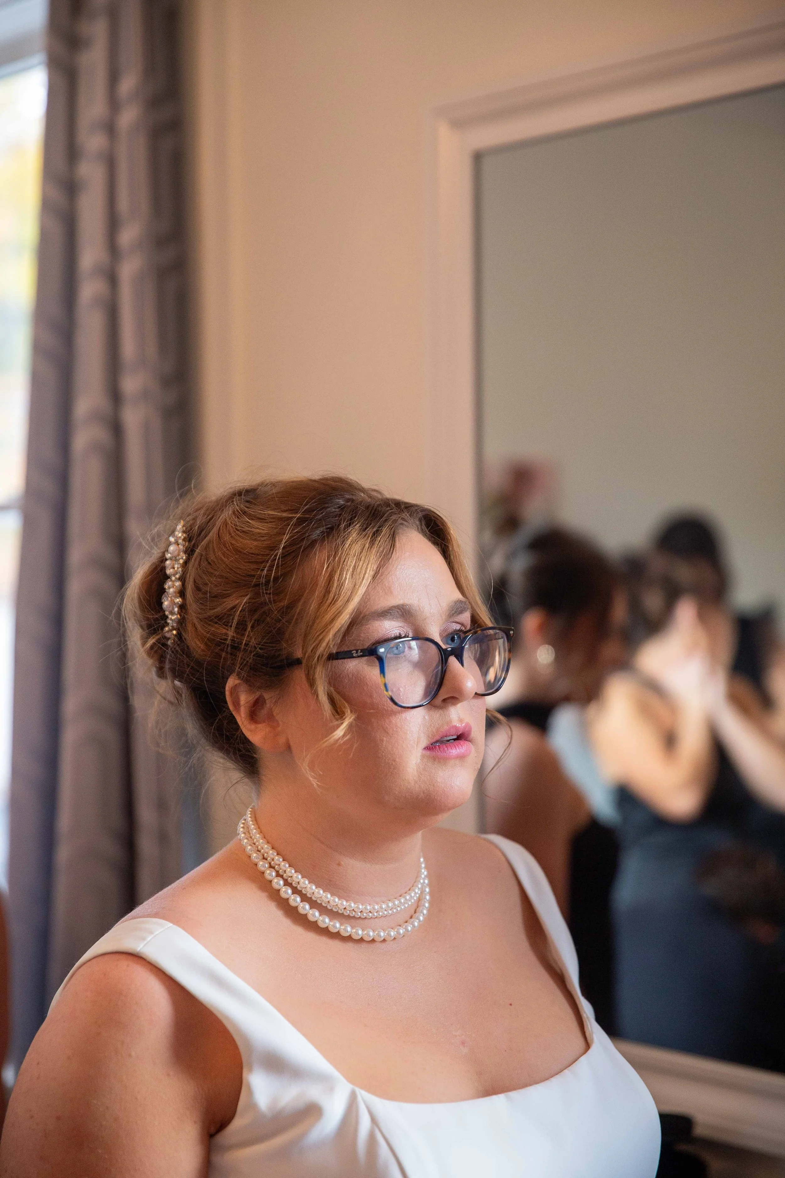 A woman with glasses and pearl jewelry looking at a mirror, with her reflection visible.