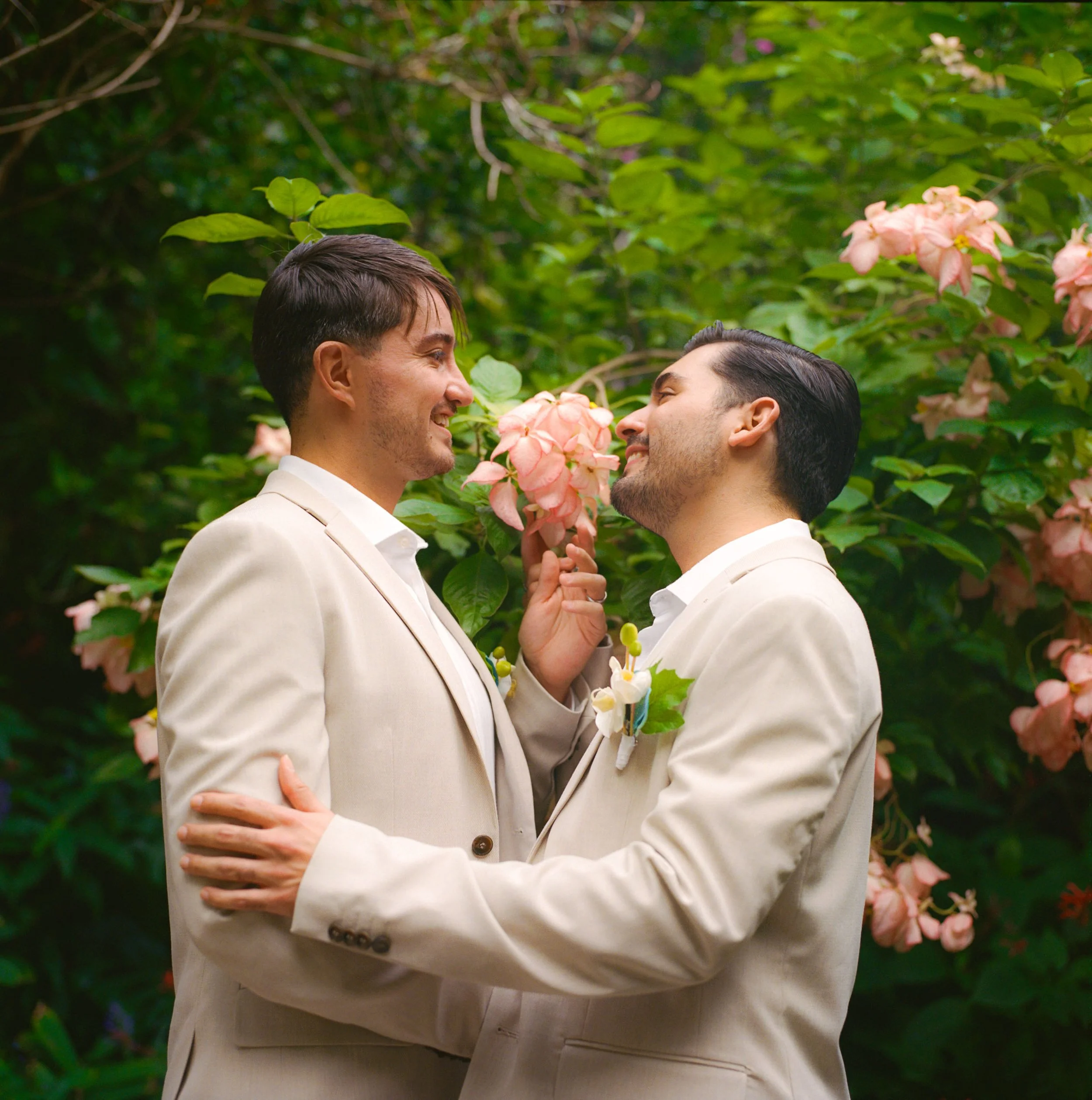 Two men in beige suits embracing outdoors with pink hydrangea flowers in the background.