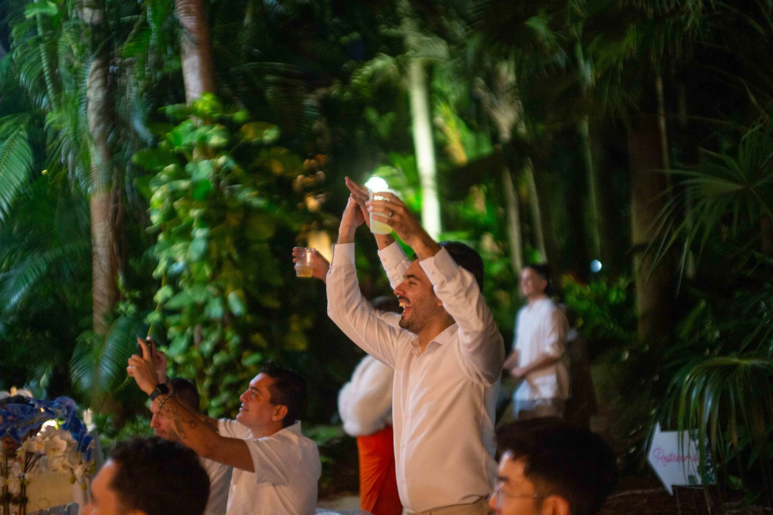 People celebrating outdoors at night surrounded by lush greenery, raising glasses and smiling.