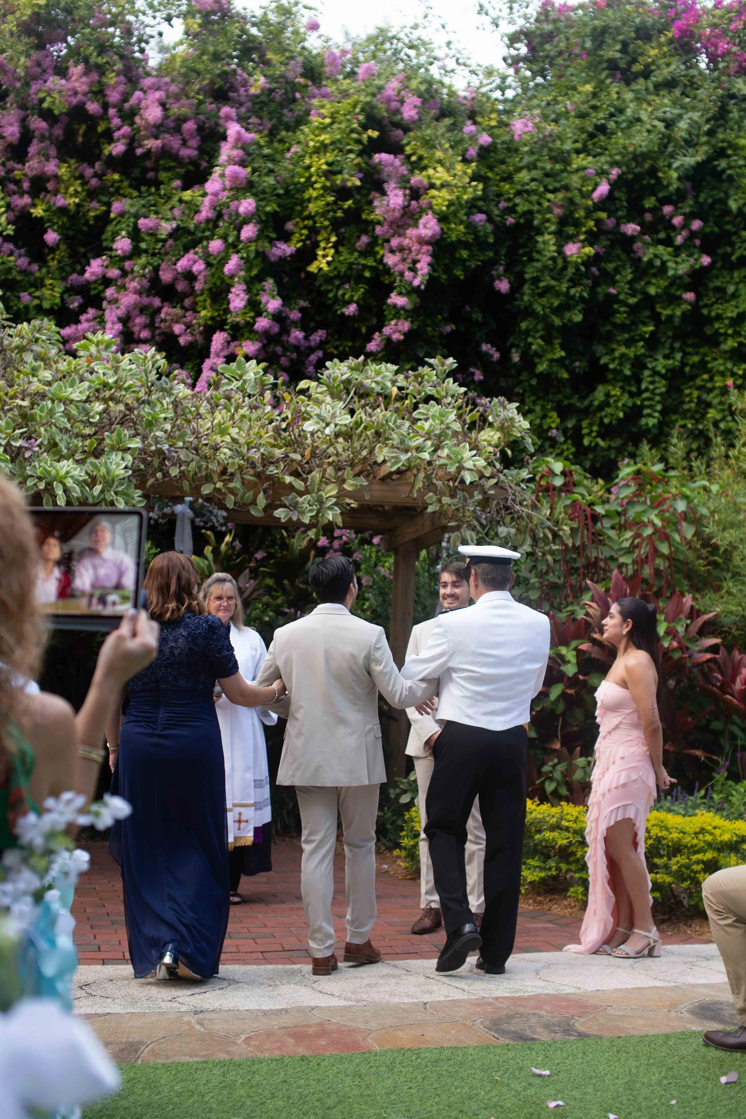 A wedding ceremony taking place outdoors in a garden with purple and pink flowers. The couple, dressed in white, is holding hands and smiling, surrounded by friends and family. A woman in a pink dress is standing nearby, and a person is taking a phot