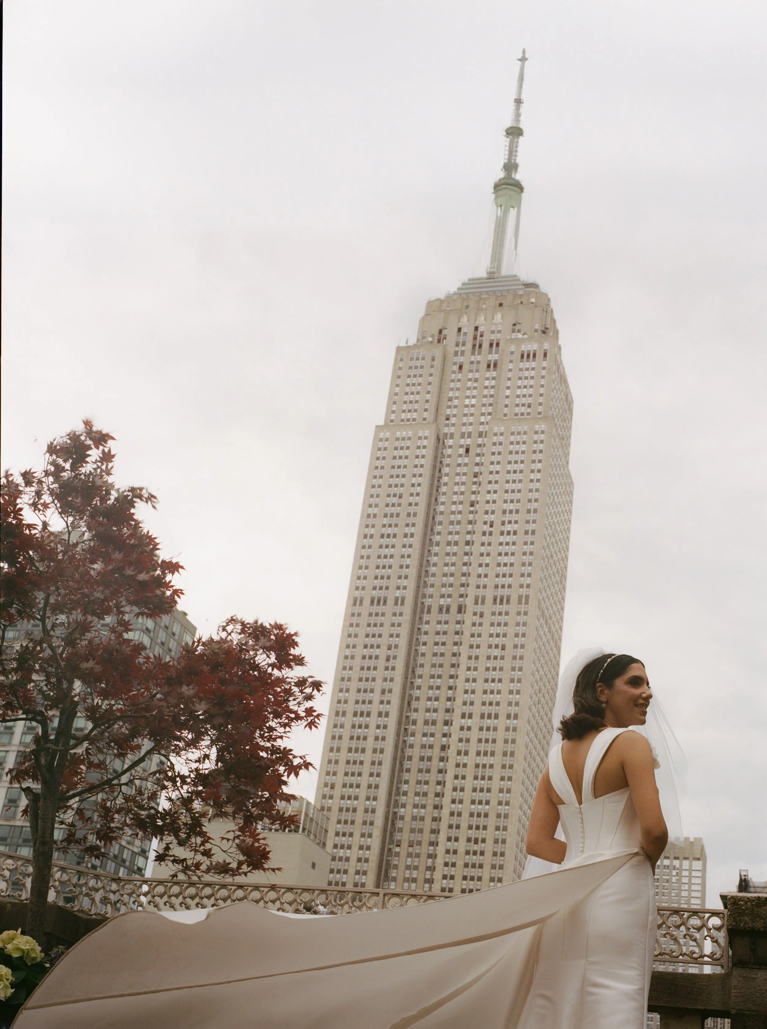Bride in white wedding dress standing outdoors with the Empire State Building in New York City in the background and cloudy sky.