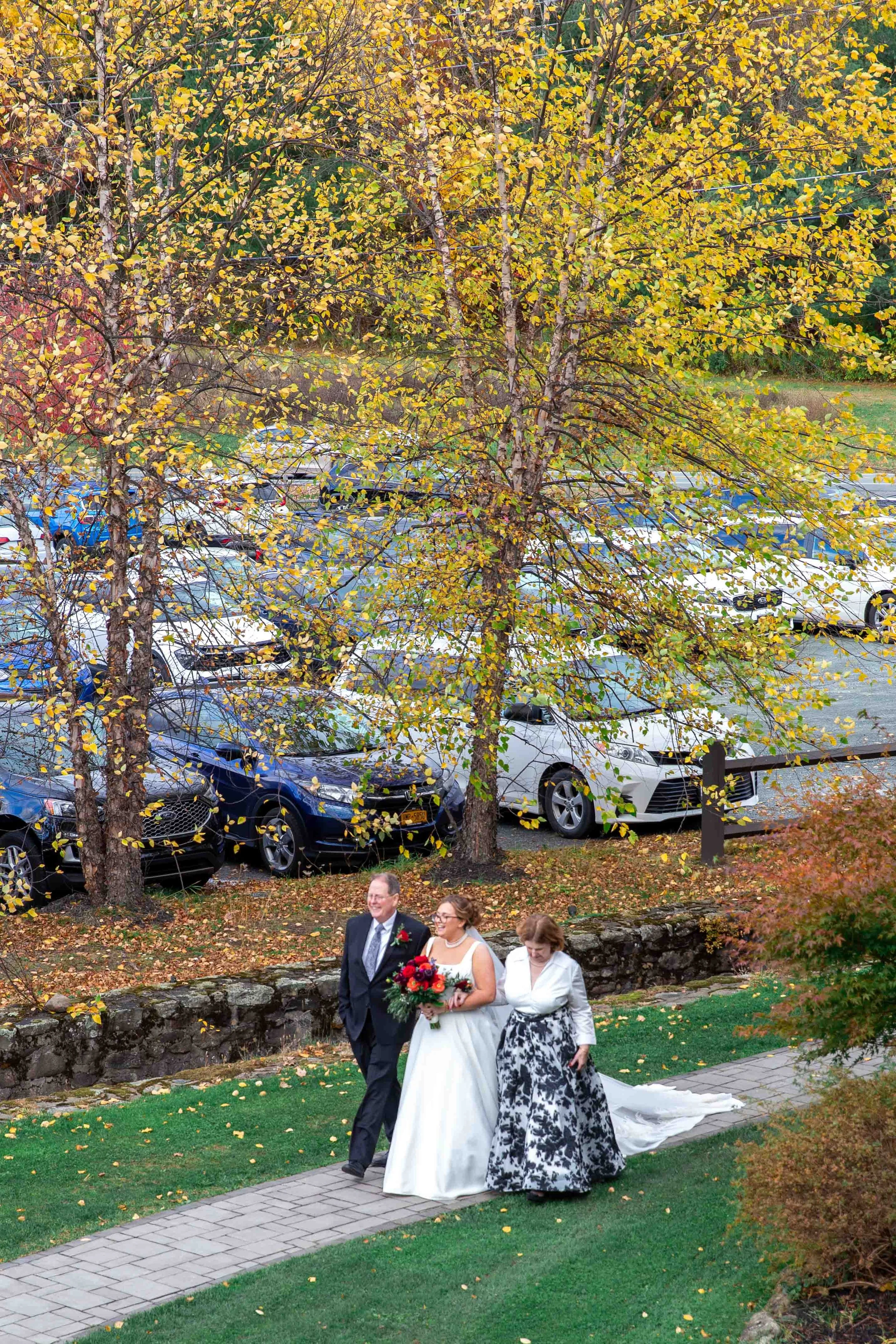 A bride in a white wedding dress holding a bouquet walking with two people, one man in a suit and one woman in a black-and-white dress, on a stone path with trees showing autumn foliage in the background.