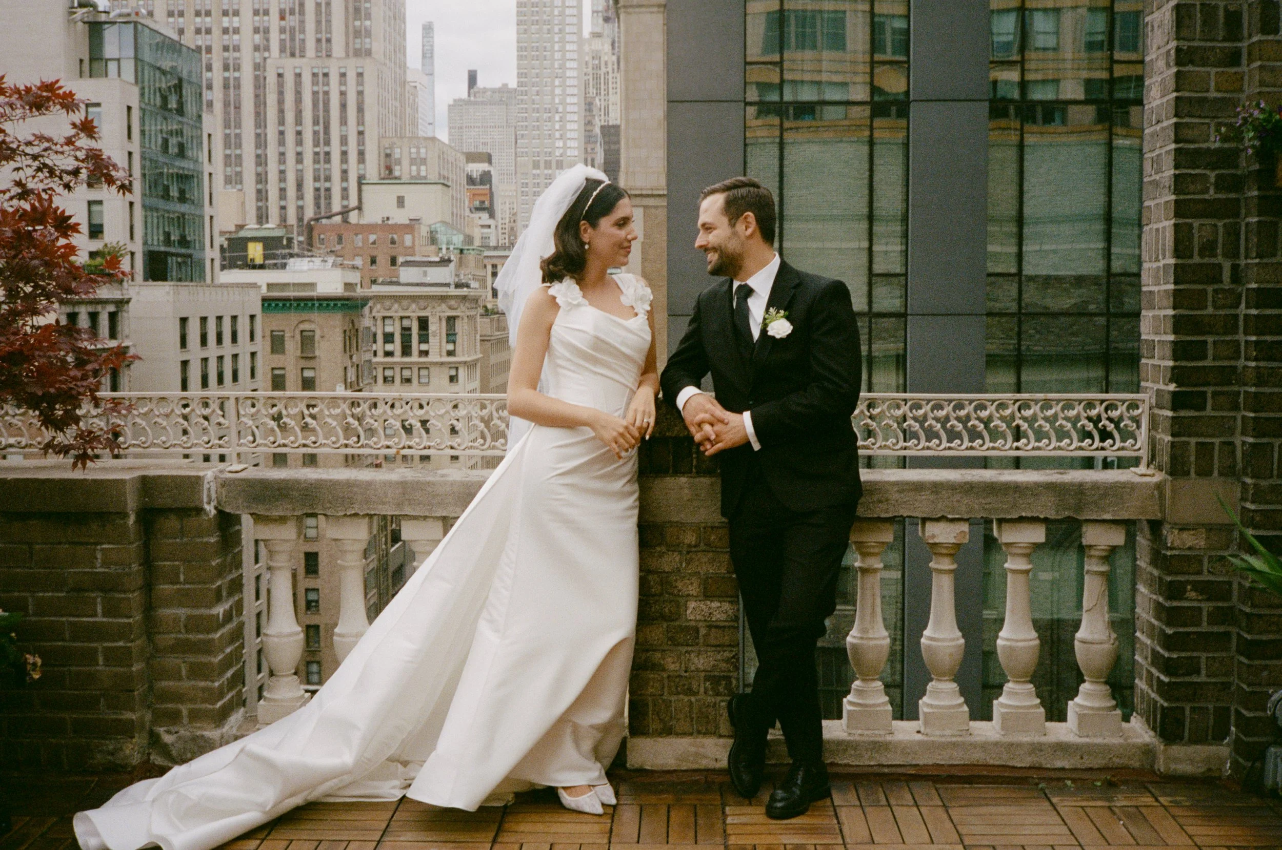 Bride and groom on a rooftop balcony in New York City, smiling at each other, with city buildings in the background.