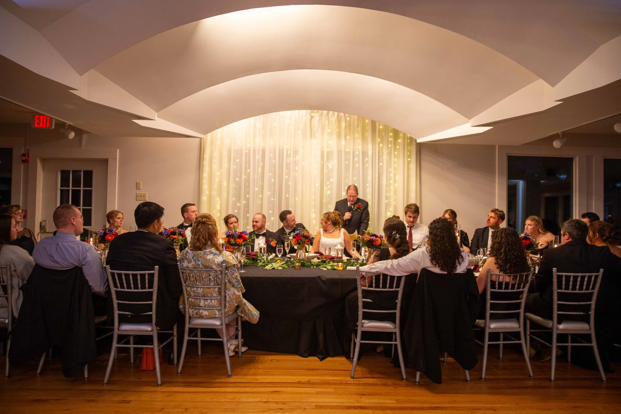 A wedding reception with guests seated around a head table, where a man is giving a speech. The background features a white curtain with string lights, and the room has curved ceiling design and hardwood flooring.