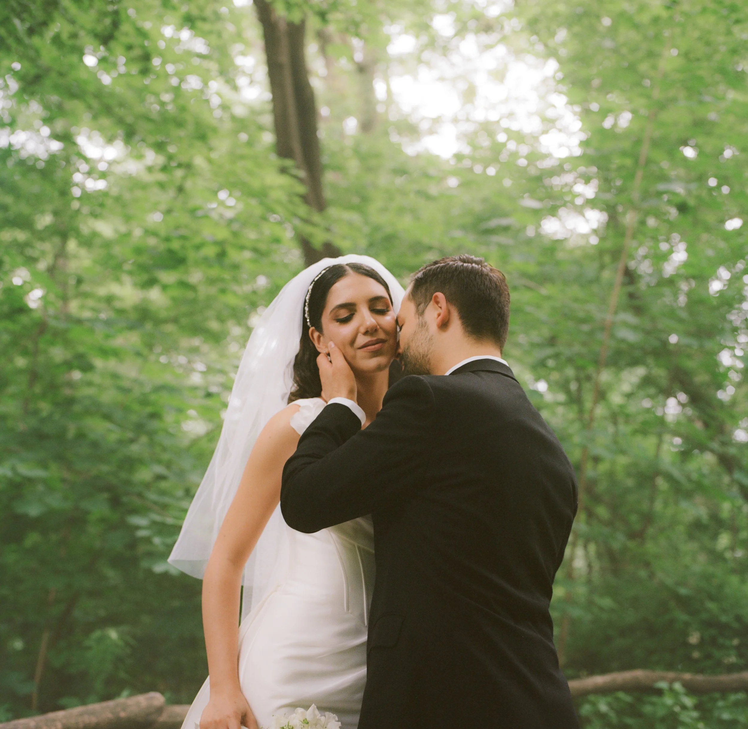 A bride in a white wedding dress with veil and a groom in a black suit embracing in a forest with green trees in the background.