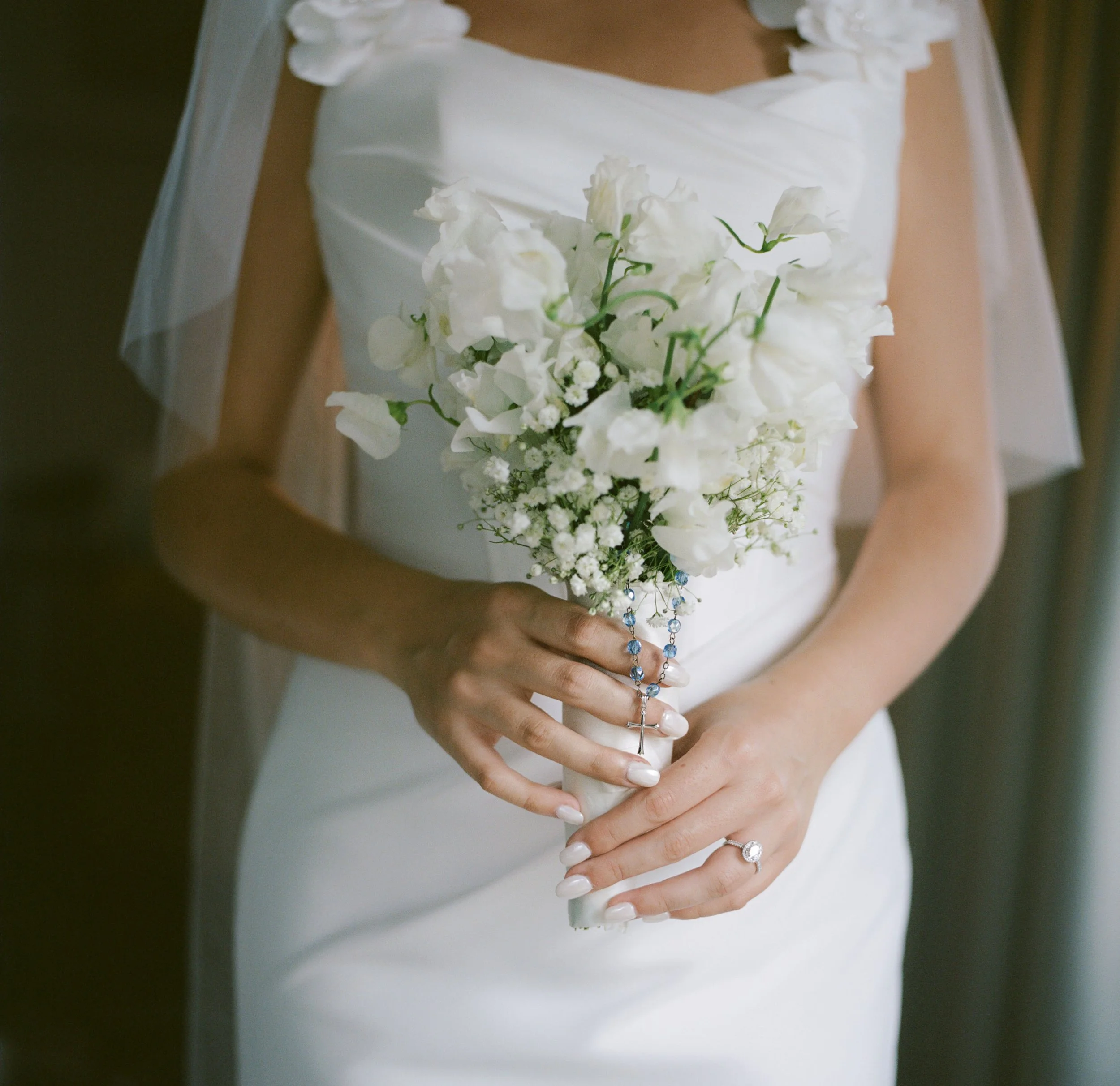 A bride holding a bouquet of white flowers and rosary beads, wearing a white wedding dress with a ring on her finger.