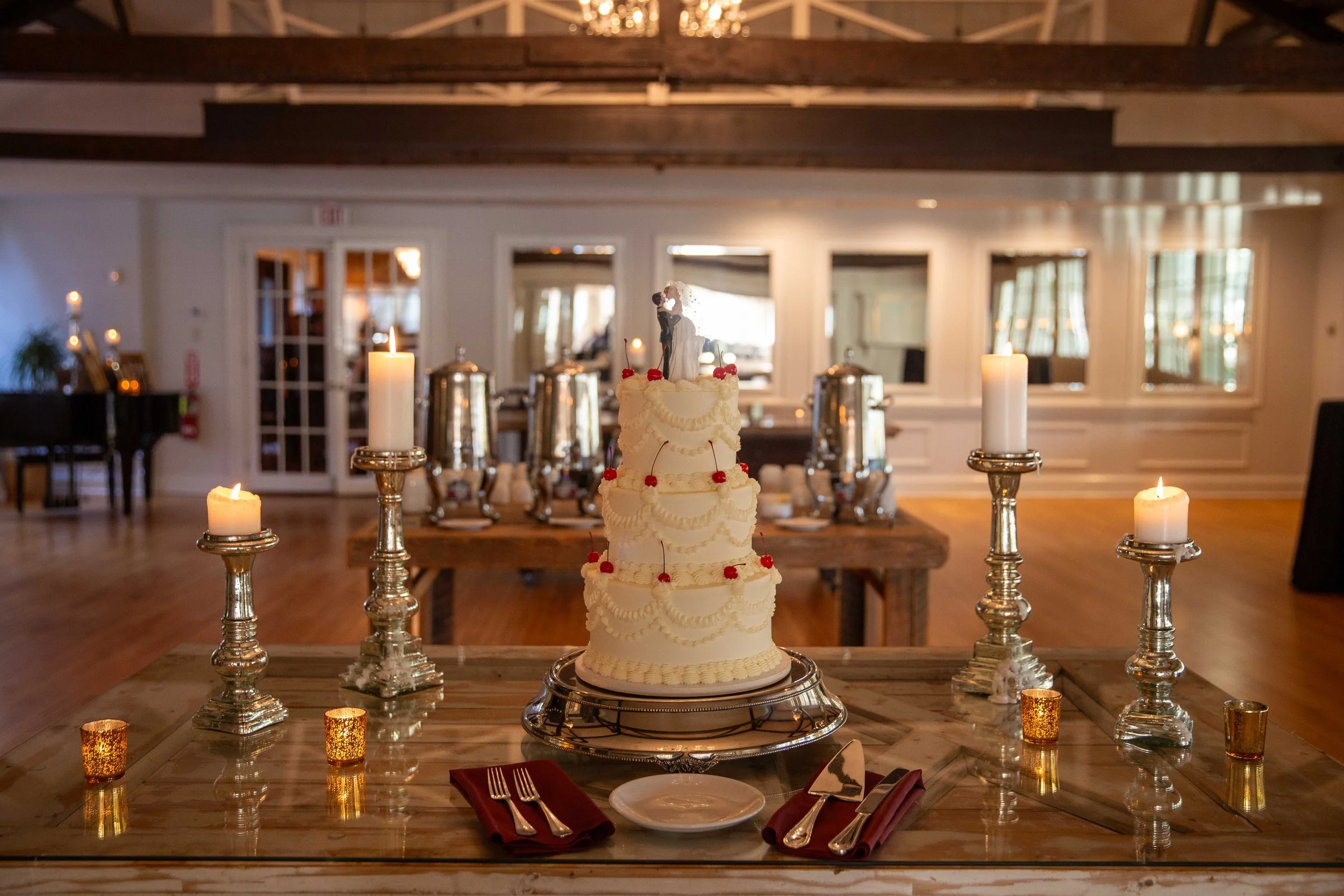 A four-tier white wedding cake with strawberries and floral decorations, surrounded by lit candles on silver candlesticks, on a glass-topped wooden table in a decorated reception hall.