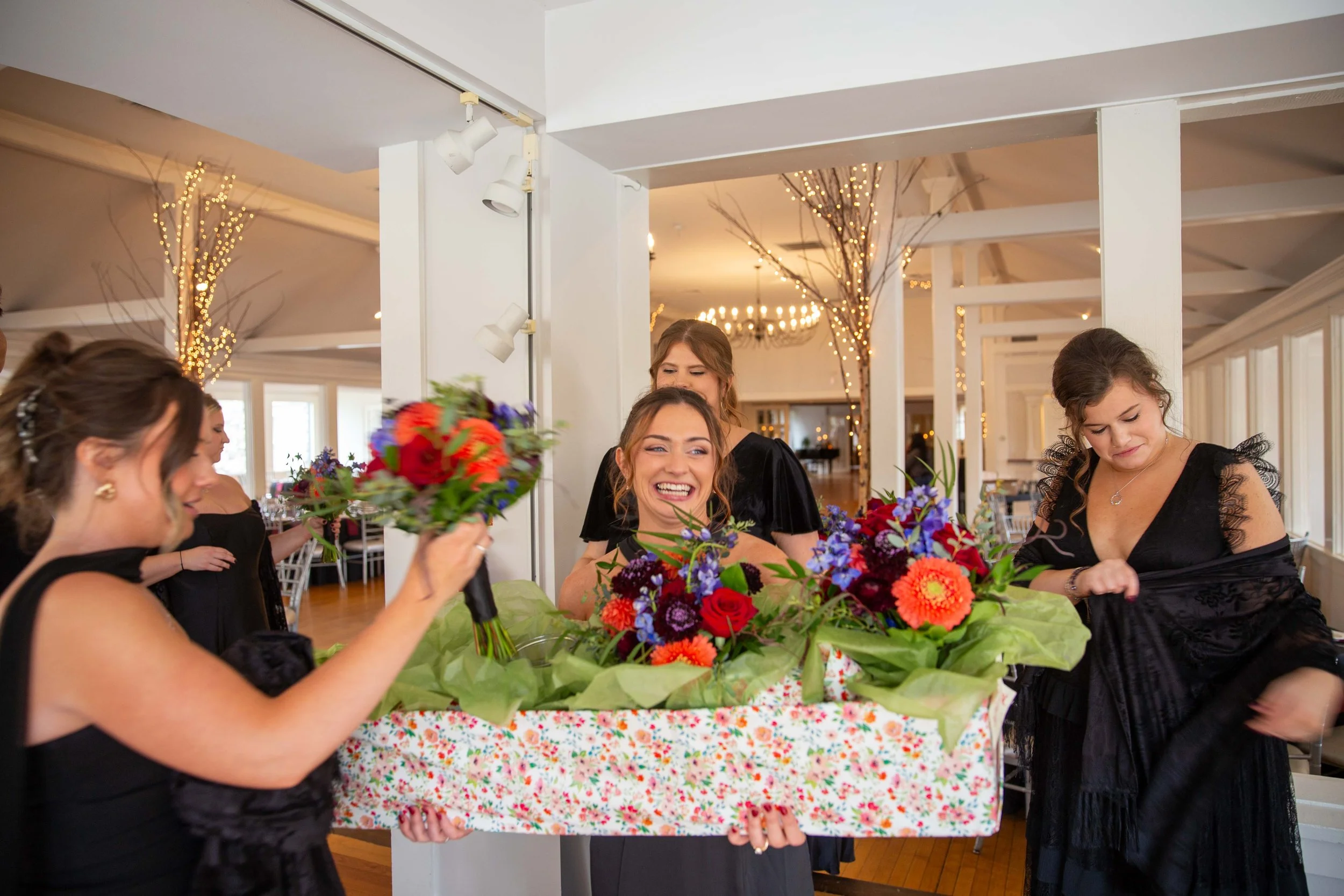 Women in black dresses at a celebration, one holding a large flower bouquet, smiling, inside a decorated venue with string lights and white walls.
