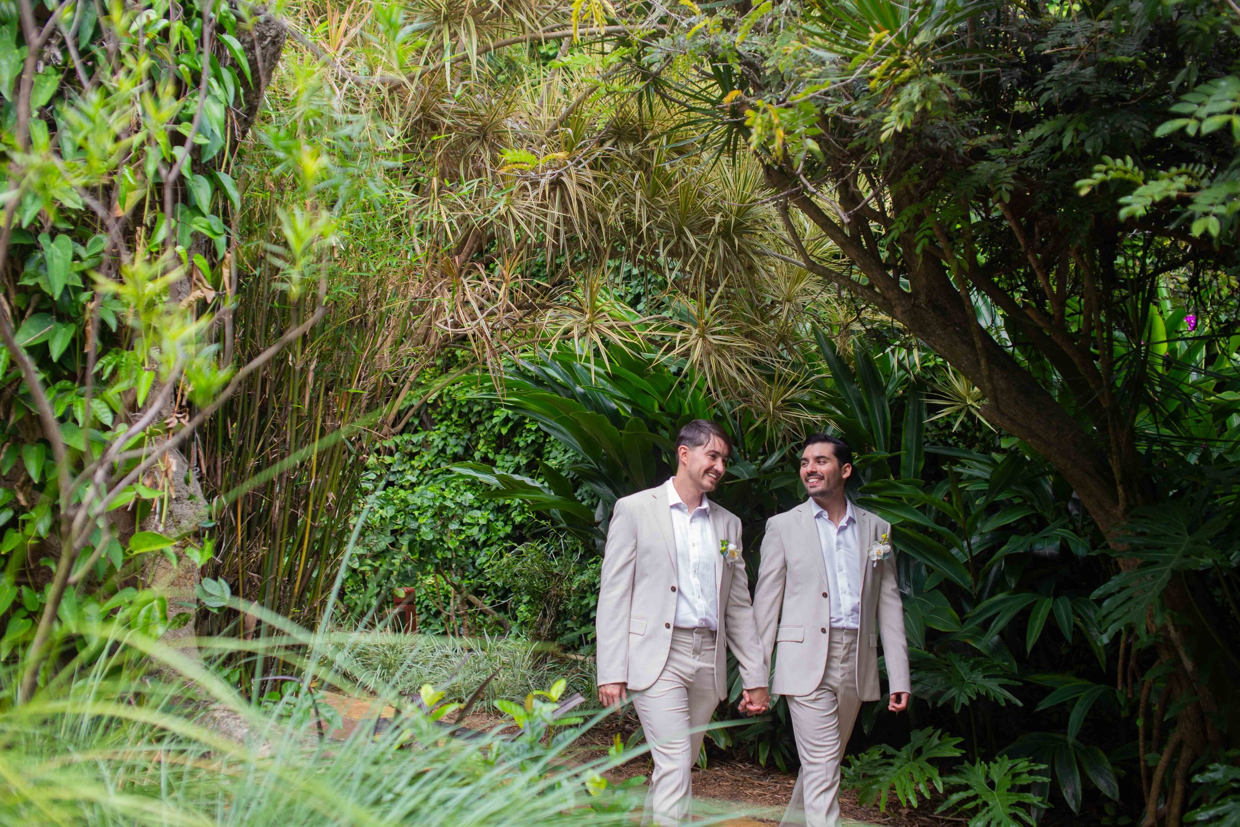 Two men dressed in light-colored suits holding hands and walking through a lush, green garden with large tropical plants and trees, smiling at each other.